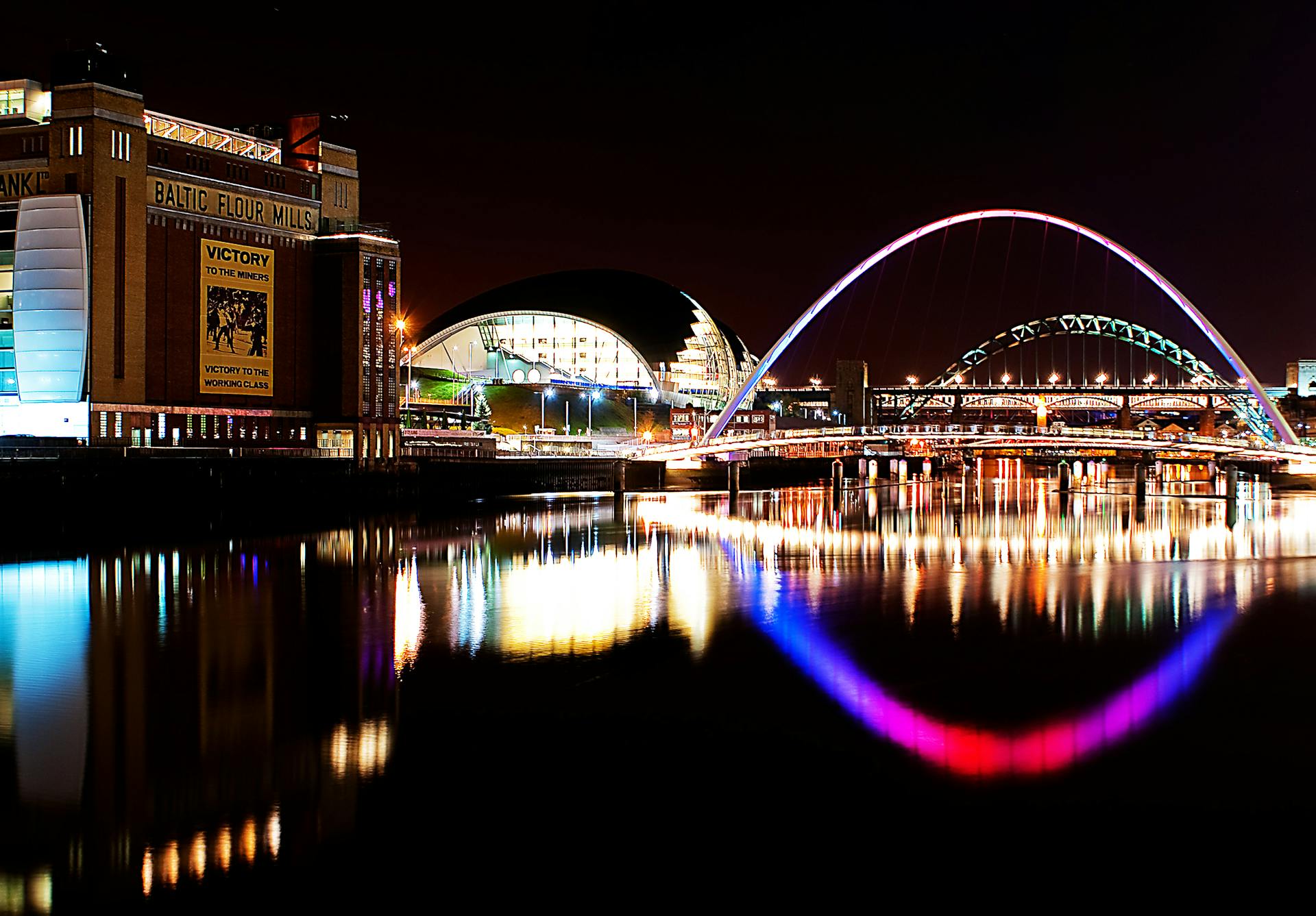 Stunning night view of Newcastle Quayside with illuminated Millennium Bridge reflecting in the Tyne River.