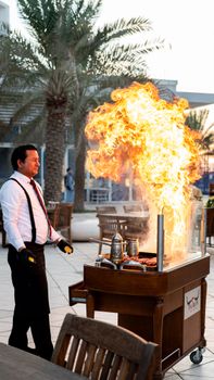 A chef creates a dramatic flambé with flames shooting high, set in a stylish outdoor restaurant.