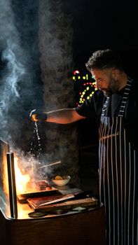 Chef expertly grilling with flames and smoke, adding seasoning for flavor at night.