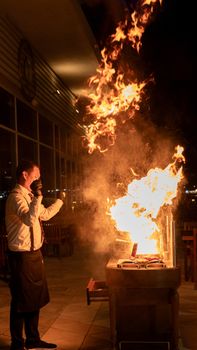 A chef performs a dramatic fiery cooking demonstration outdoors at night.