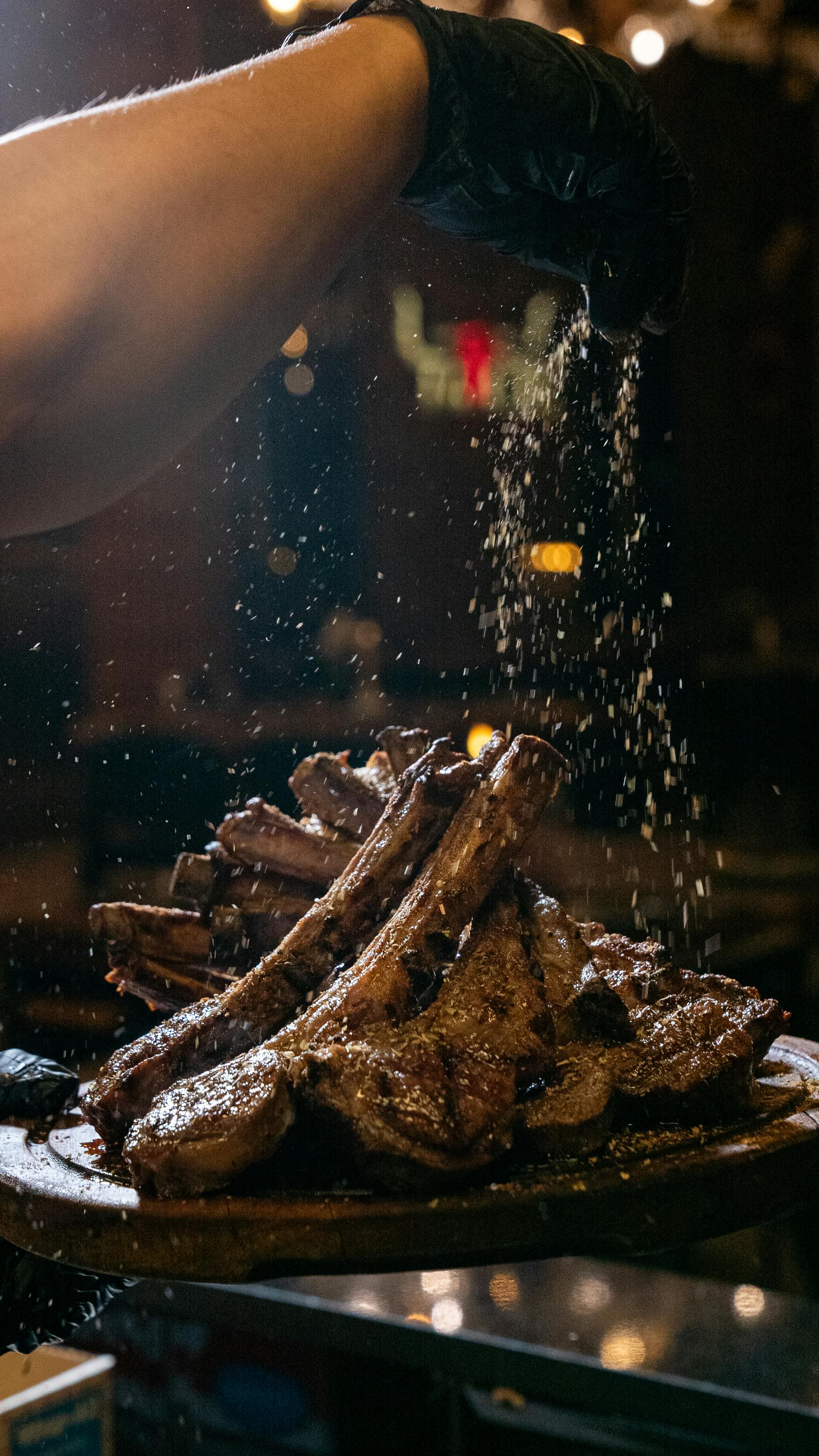 grátis Close-up de um chef temperando costeletas de cordeiro grelhadas com ervas em uma cozinha rústica com pouca luz. Foto profissional