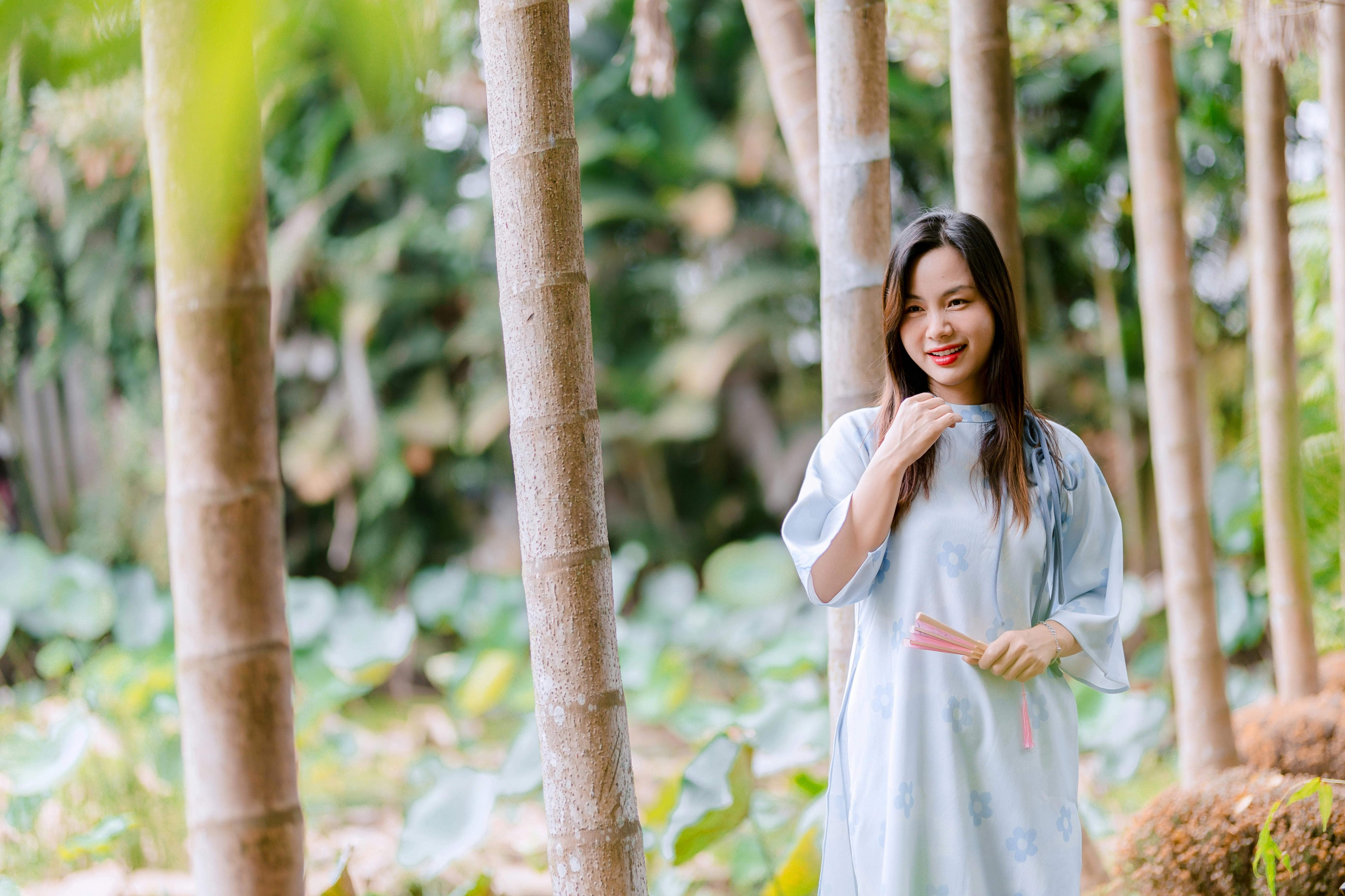 gratis Een jonge vrouw in een lichtblauwe jurk staat glimlachend tussen de bomen in een weelderige tuin. Stockfoto