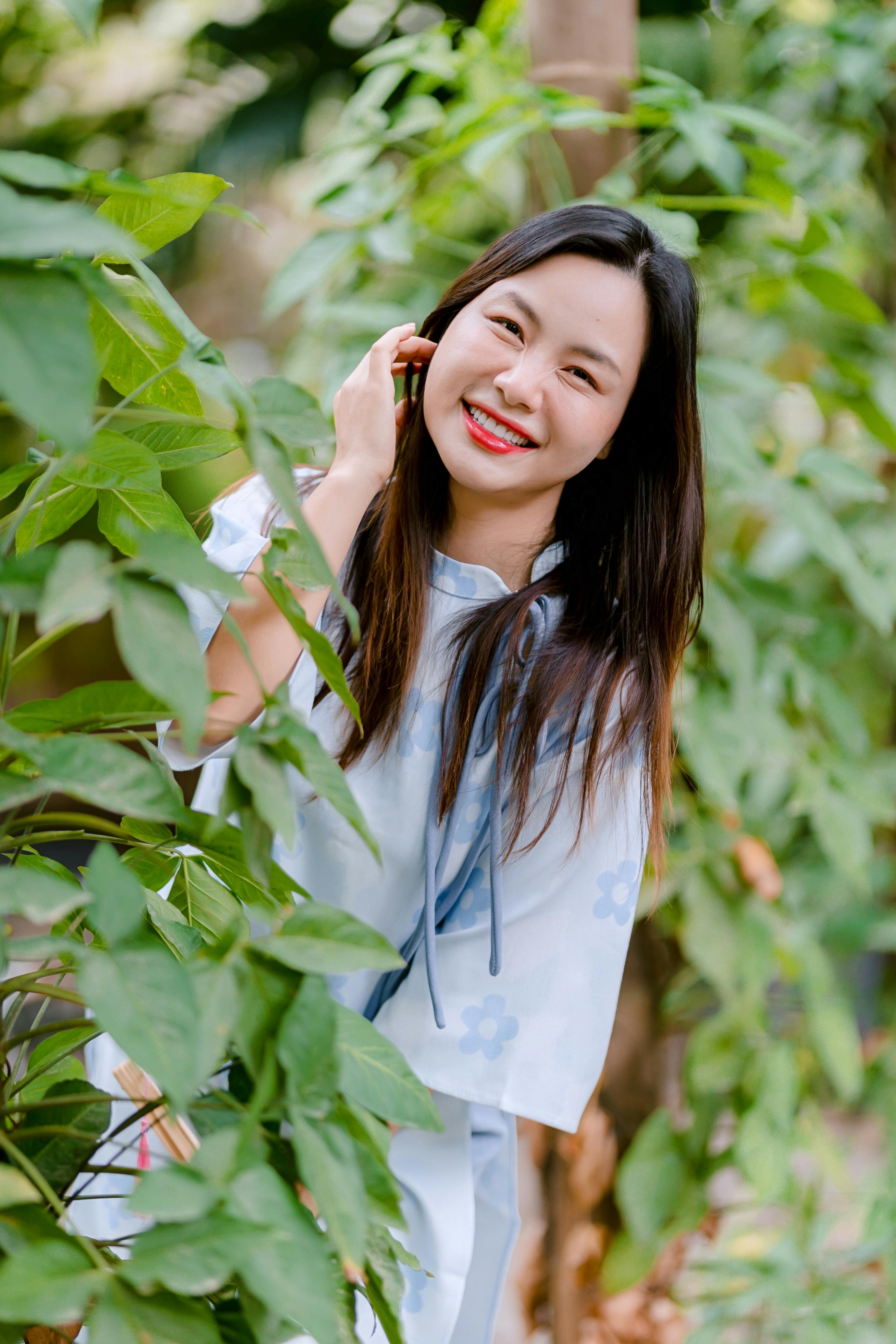 Gratis En munter kvinde i en blå kjole står blandt livlige grønne planter og smiler bredt udendørs. Lagerfoto