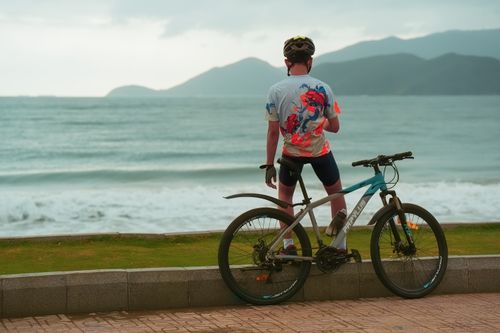 Free A cyclist pauses on a coastal pathway, gazing at the sea with a scenic mountain backdrop. Stock Photo