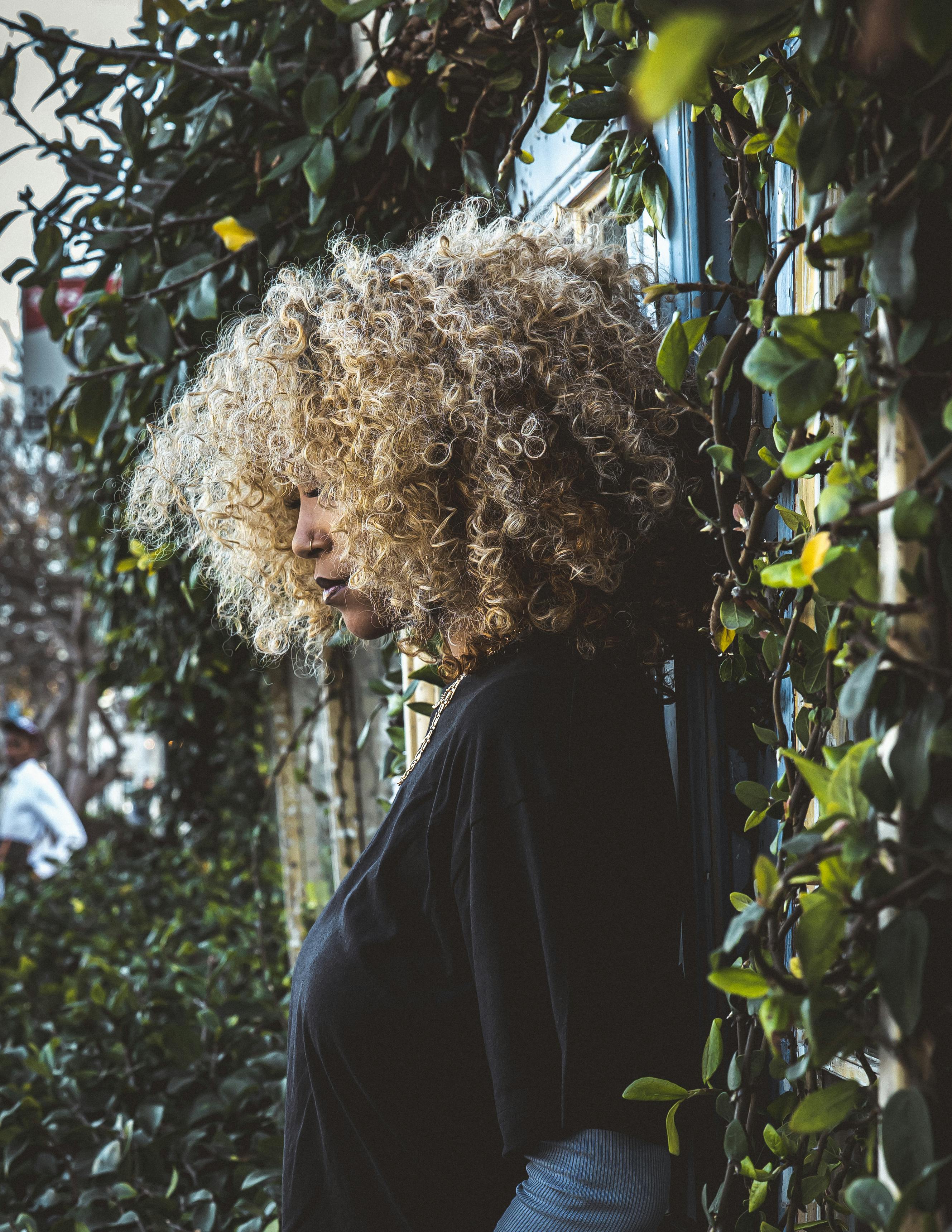 gratis Een vrouw met krullend haar staat bij een met planten begroeide muur, verdiept in gedachten. Stockfoto