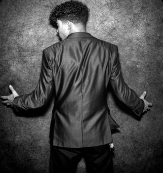 A young man in a black suit facing a textured wall, captured from behind in black and white.