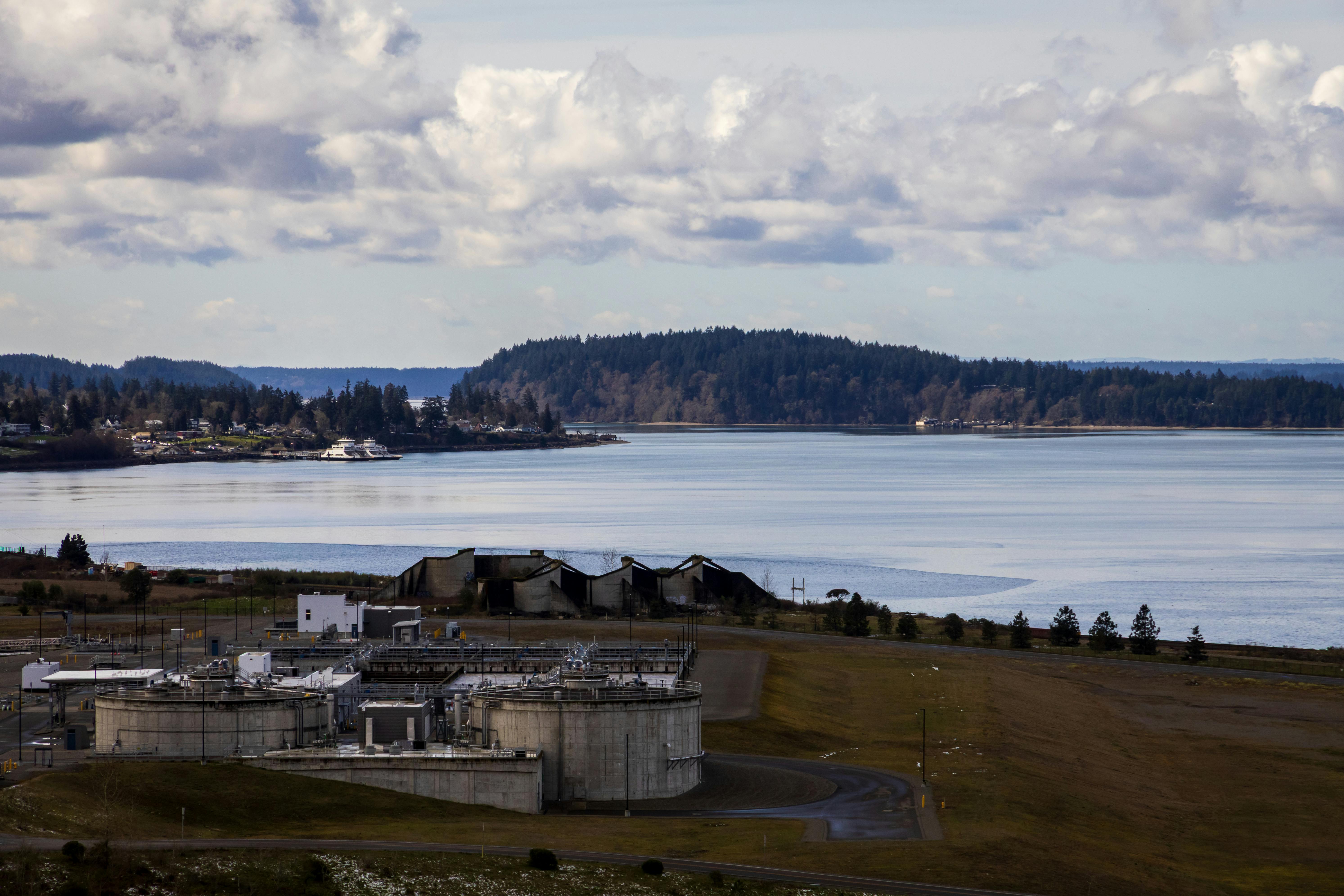 бесплатная Промышленный комплекс расположен недалеко от спокойной набережной, окруженной лесистыми холмами и облаками, что создает контраст между природой и индустриальным ландшафтом. Стоковое фото