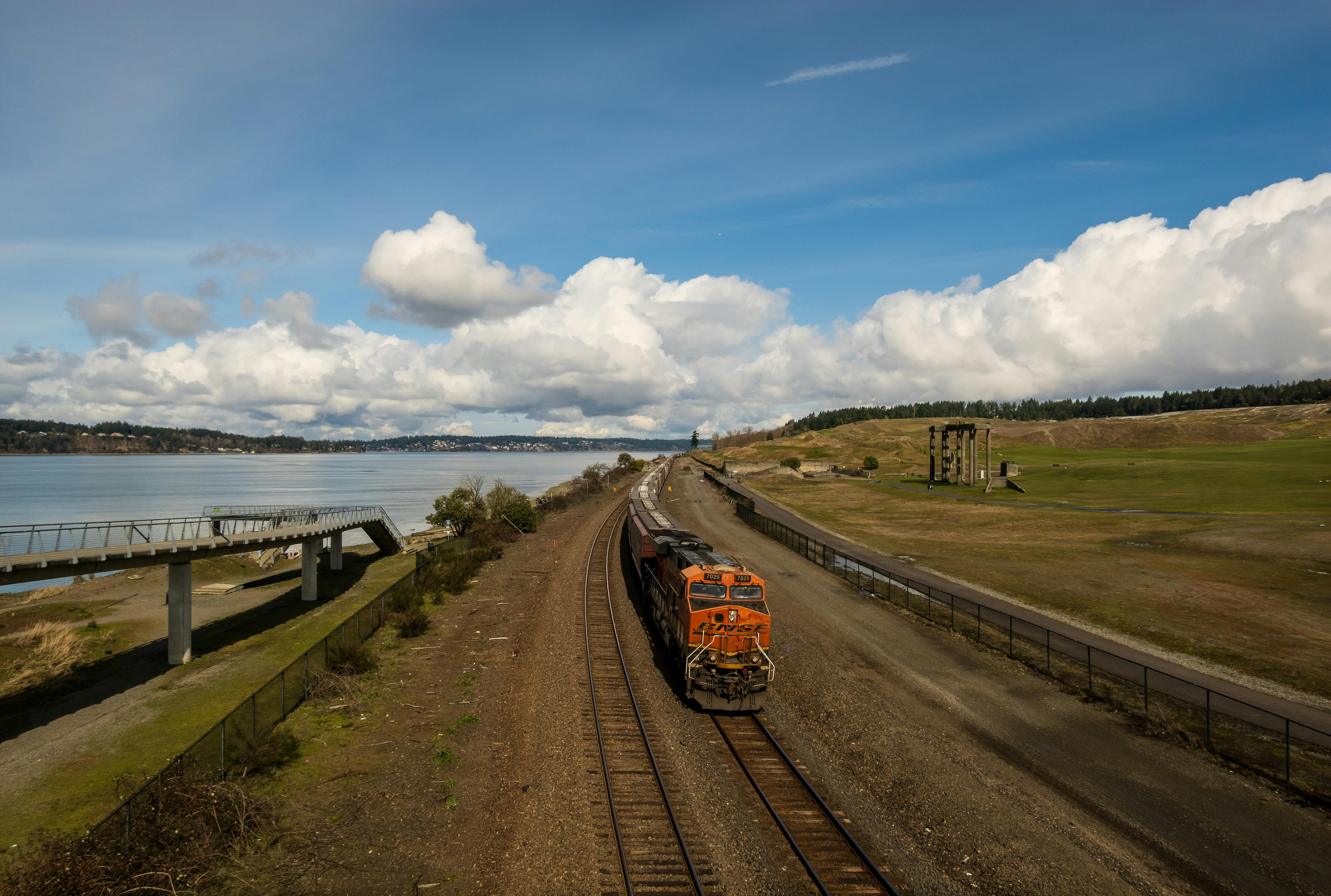 Gratis Et fargerikt godstog kjører langs en jernbane ved innsjøen med frodige, grønne marker under en delvis overskyet himmel som bakgrunn. Arkivbilde