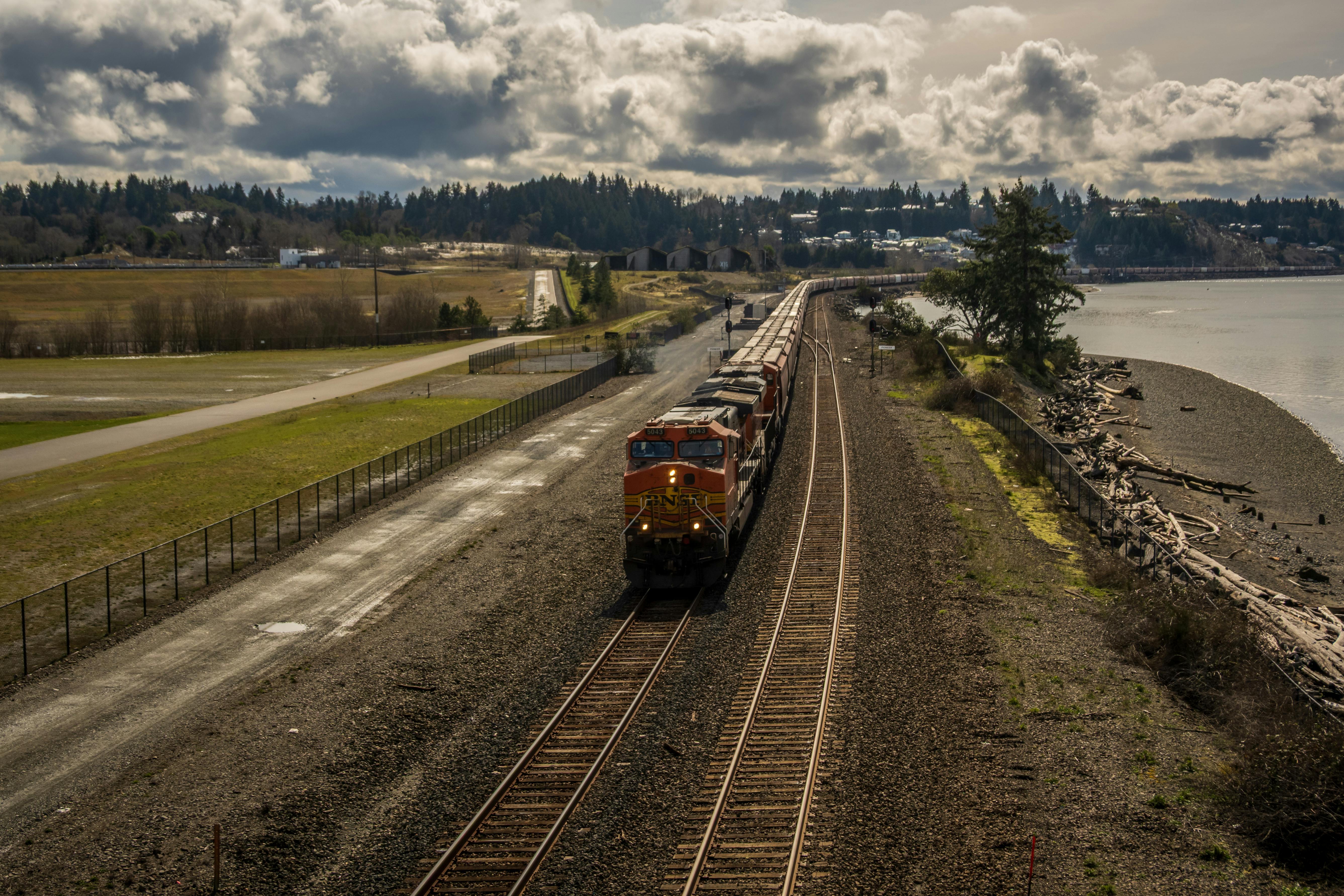 De franc Un tren de mercaderies taronja viatja per un paisatge exuberant vora l'aigua, destacant el transport i la natura. Foto d'estoc