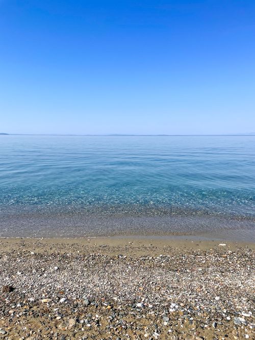 Free Serene view of a pebbled beach meeting the crystal-clear blue sea on a sunny day. Stock Photo