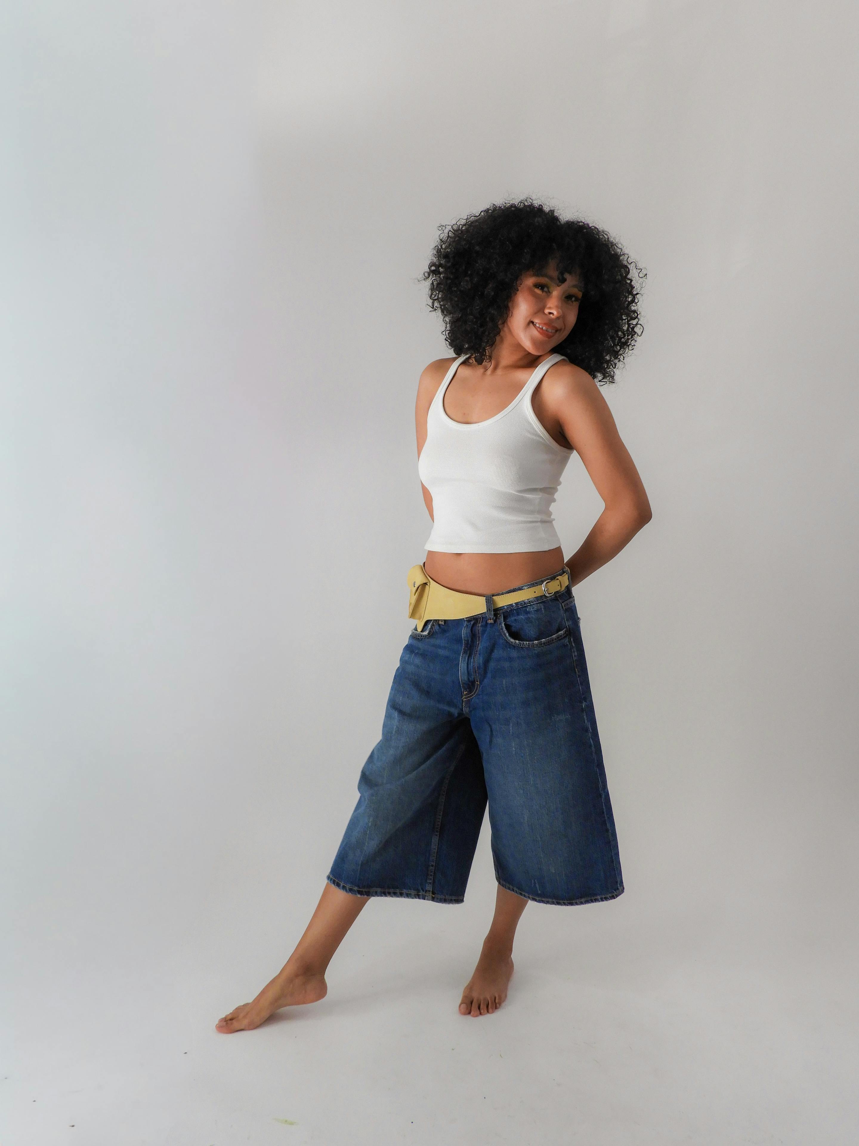 Gratis Stilig portrett av en kvinne i denimshorts og hvit tanktop som poserer selvsikkert mot en nøytral bakgrunn. Arkivbilde