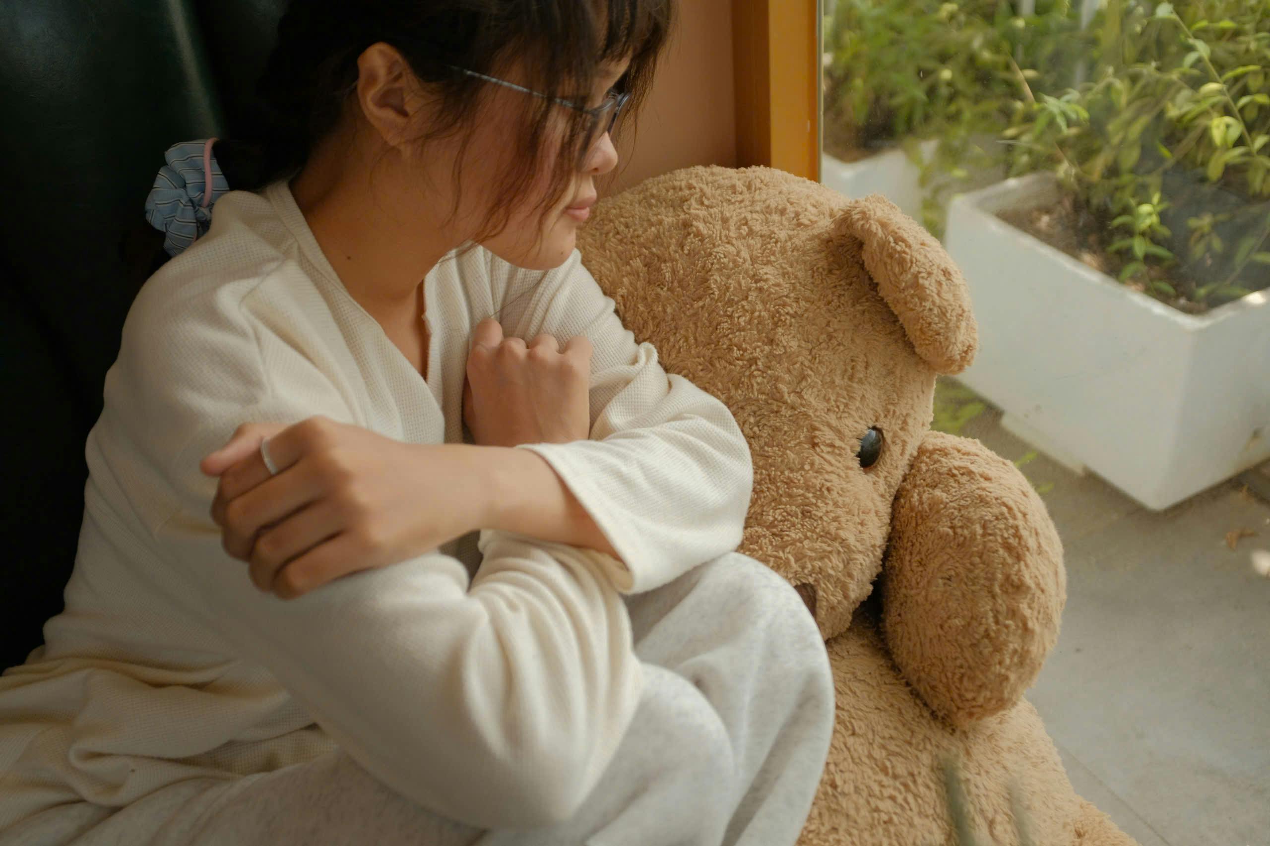 무료 흰색 잠옷을 입은 아시아 여성이 실내 창가에서 곰인형을 껴안고 있다. 스톡 사진