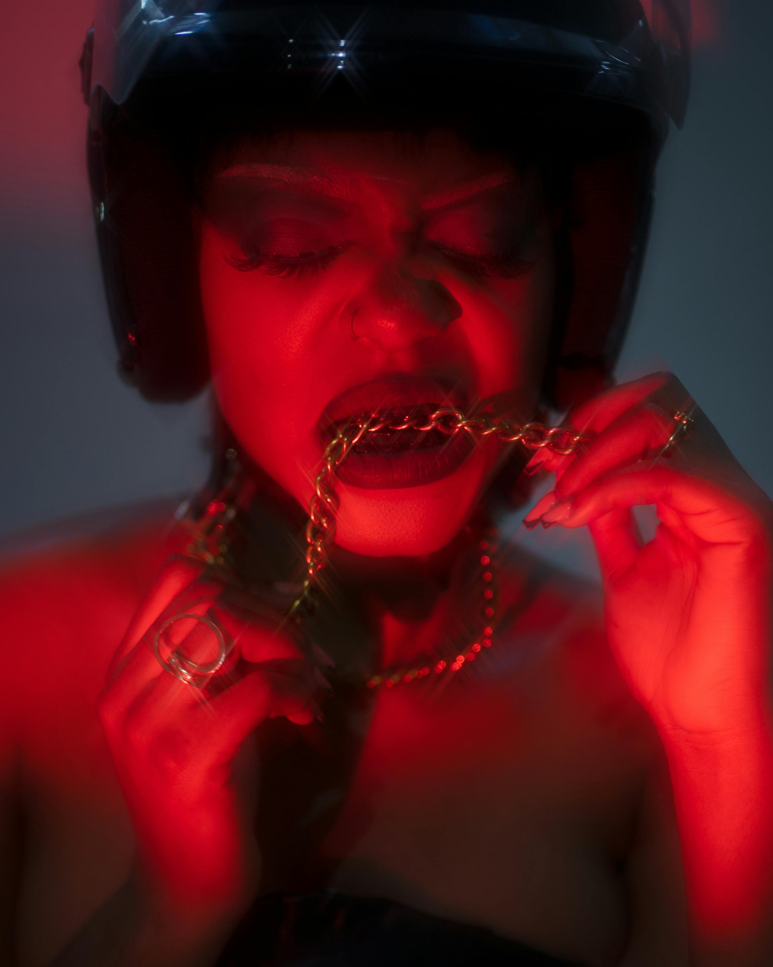 Gratis Stiliseret portræt med dramatisk belysning, der viser en kvinde, der bider i en guldkæde, iført hjelm. Lagerfoto