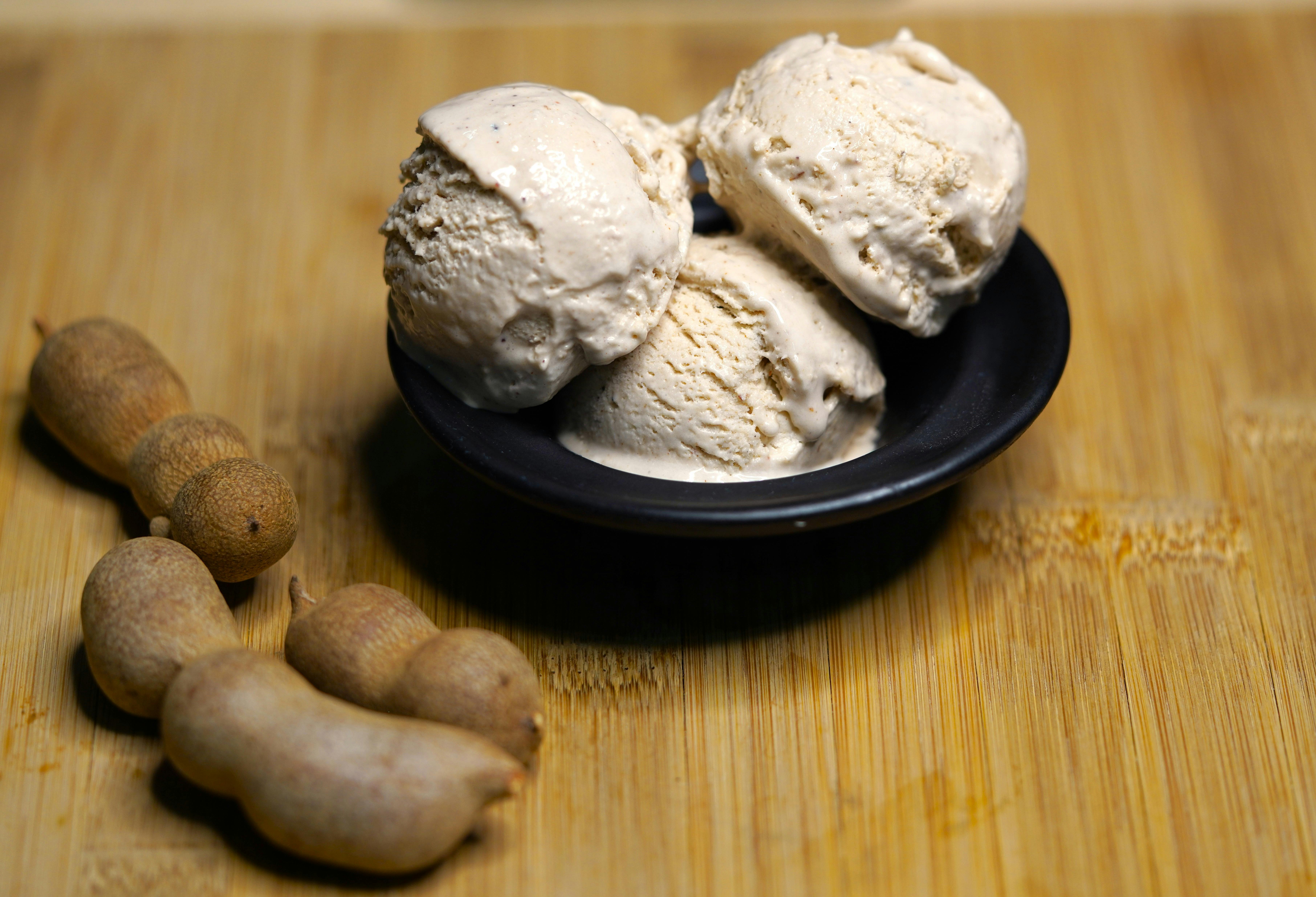 Bezpłatne Gałki kremowych lodów tamaryndowych w czarnej misce na drewnianej desce, obok strąków tamaryndowca. Zdjęcie z galerii