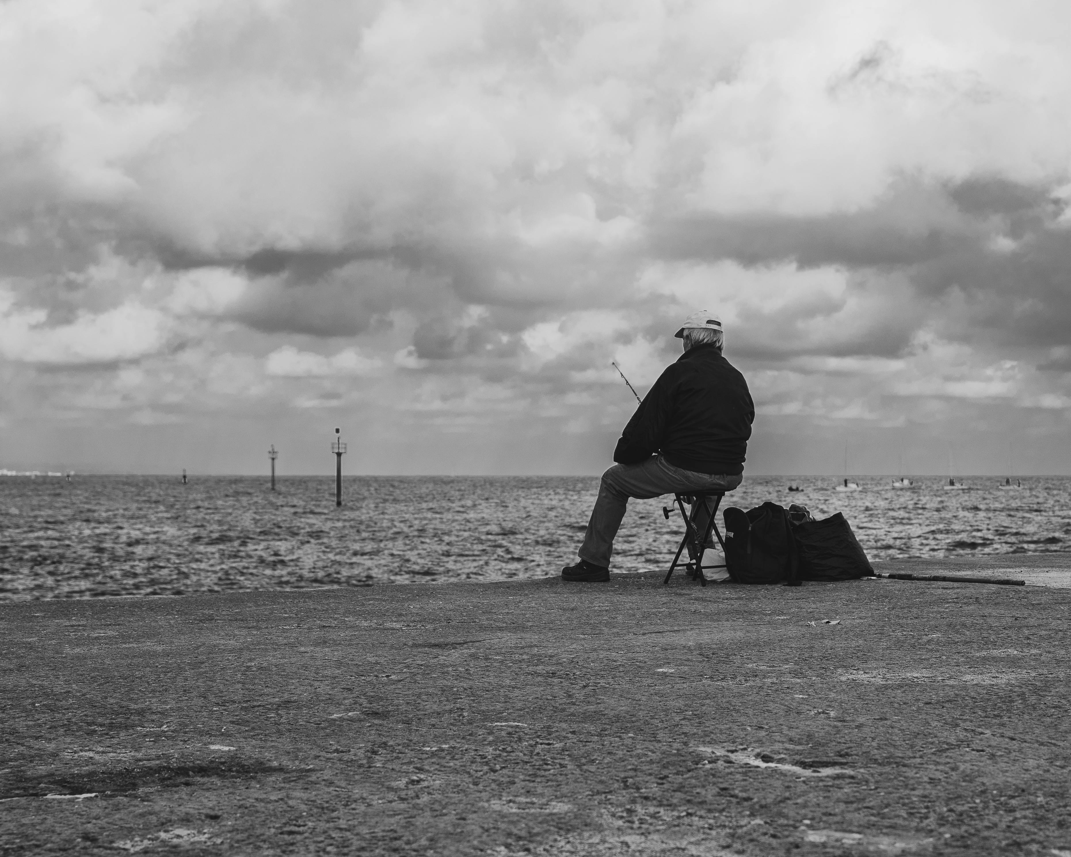 Gratis Imagen monocromática de un pescador solitario sentado junto al mar en España, que irradia tranquilidad. Foto de stock