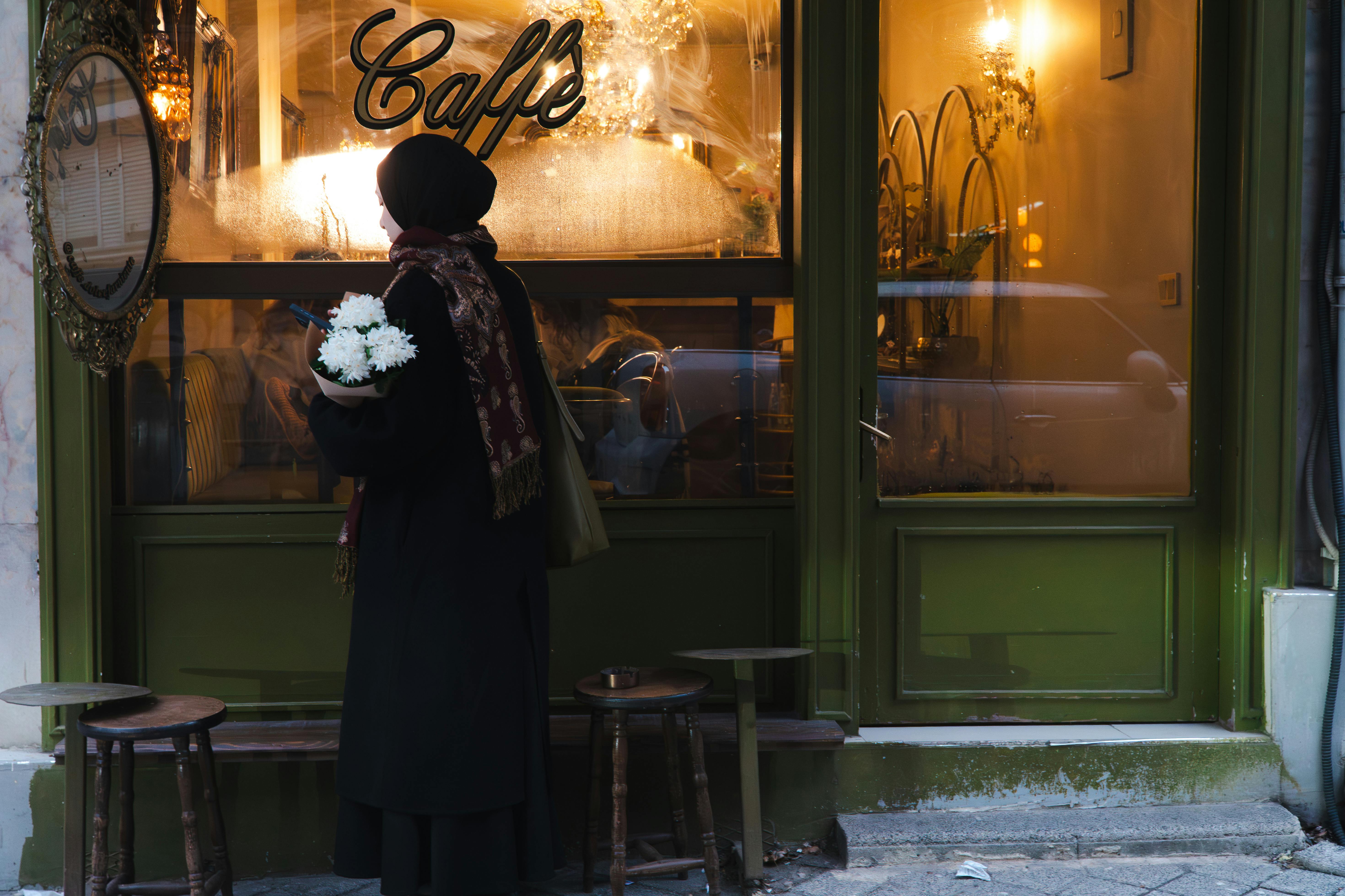Gratuit O femeie ținând flori în mână stă în fața unei cafenele confortabile și călduros luminate din Istanbul. Fotografie de stoc