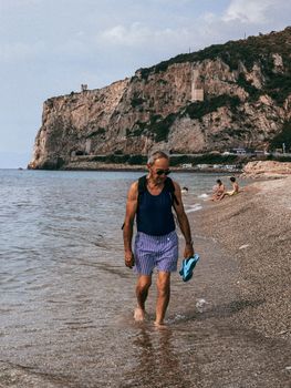 An elderly man strolls along a picturesque Italian beach with towering cliffs in the background.