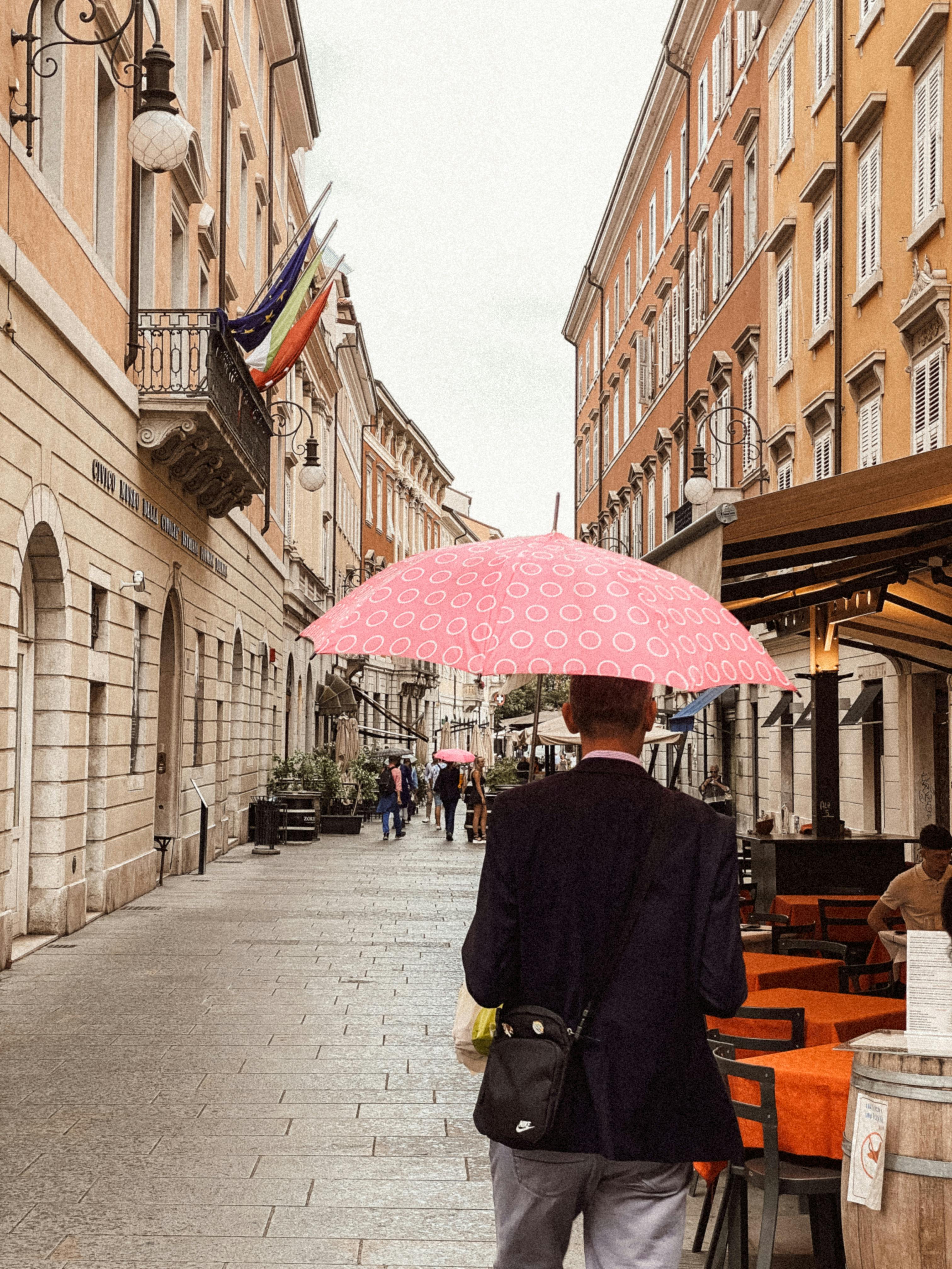 Gratis En mand med en lyserød paraply går ned ad en charmerende gade i Italien og indfanger essensen af ​​italiensk arkitektur. Lagerfoto