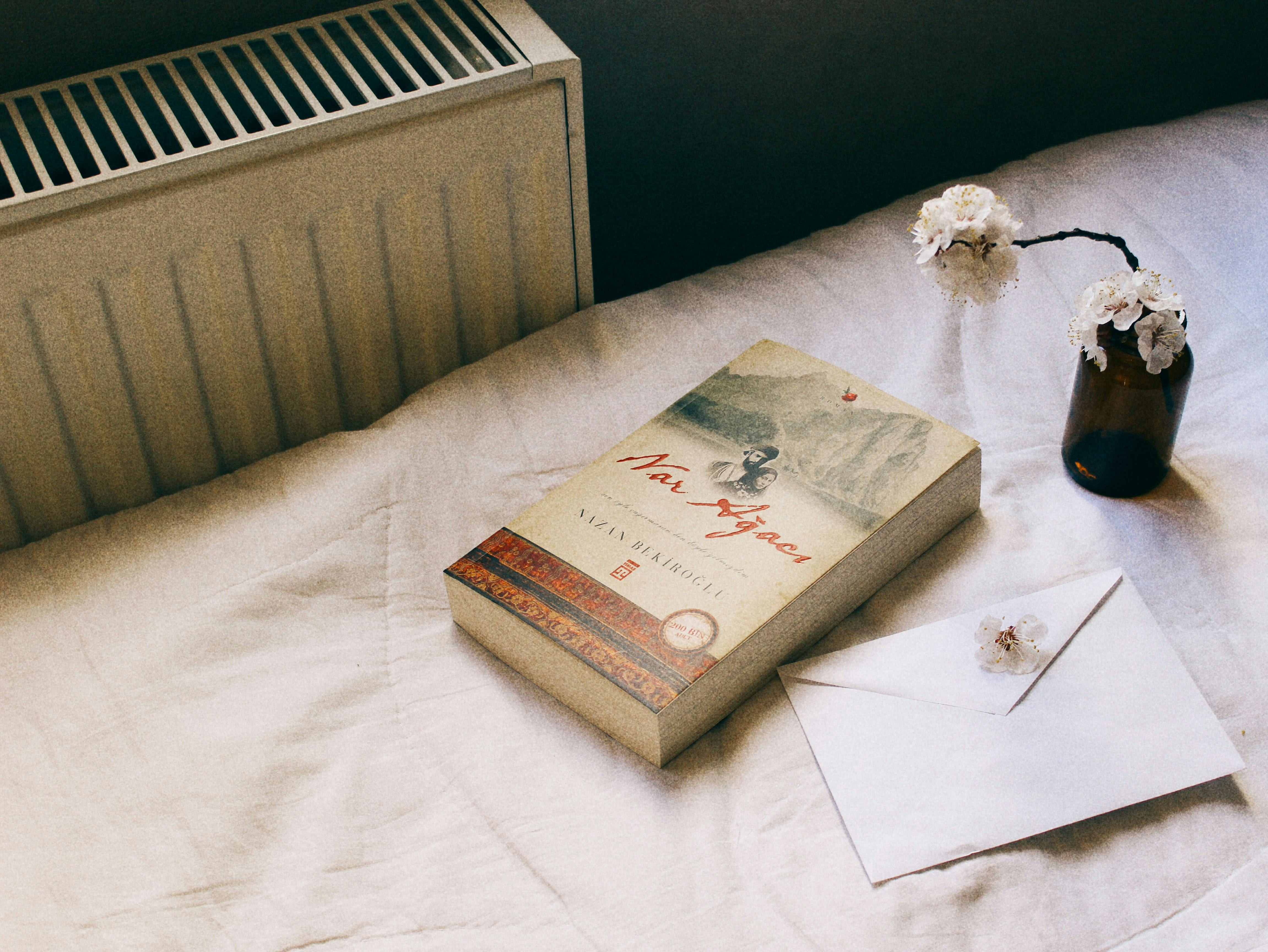 gratis Een uitnodigende afbeelding van een boek, een vaas met bloemen en een envelop op een knus bed, ideaal voor thema's rond ontspanning. Stockfoto