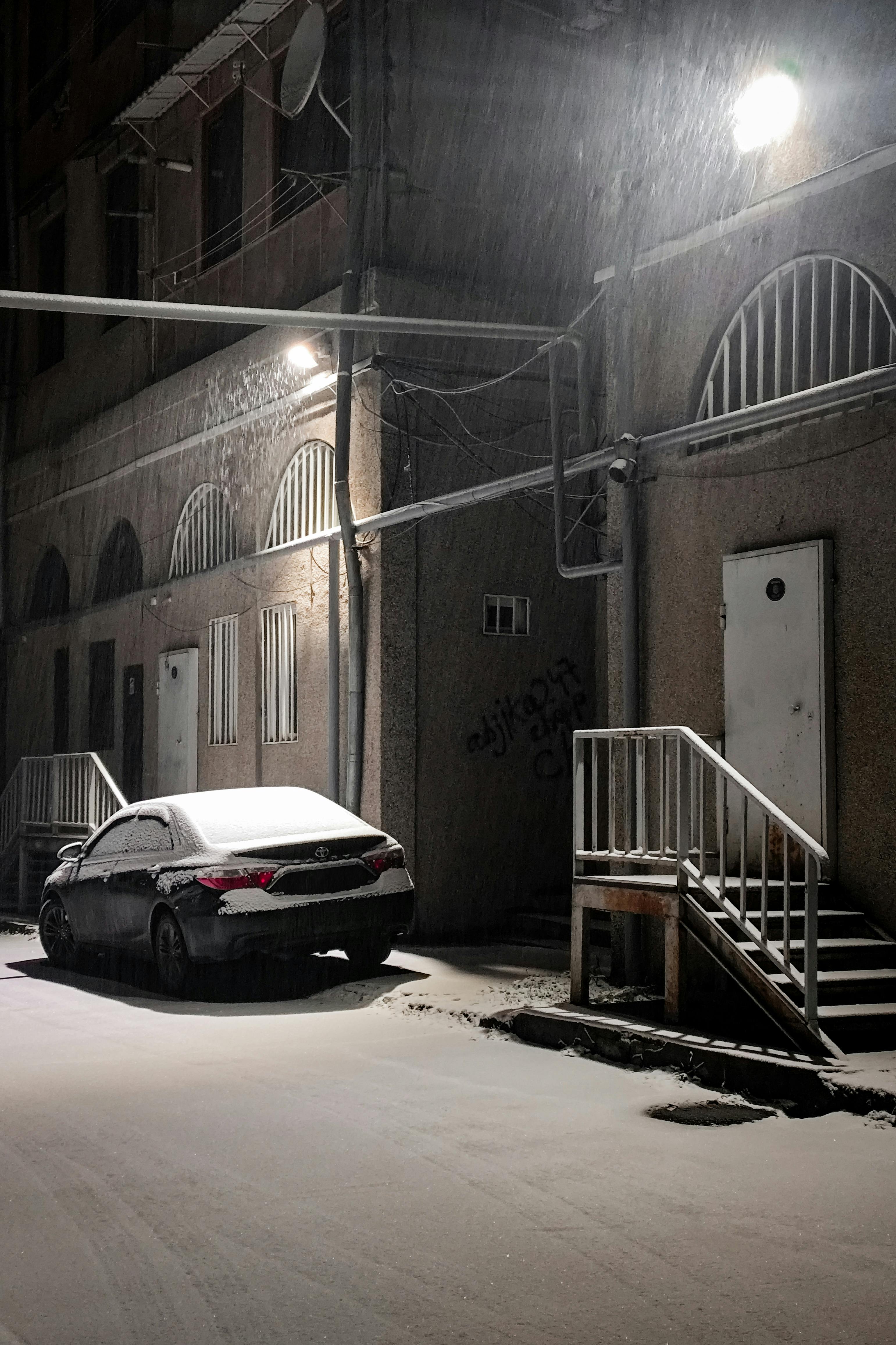 gratis Een stille stadsstraat bedekt met sneeuw, met een geparkeerde auto en een verlichte gebouwgevel in de koude nacht. Stockfoto