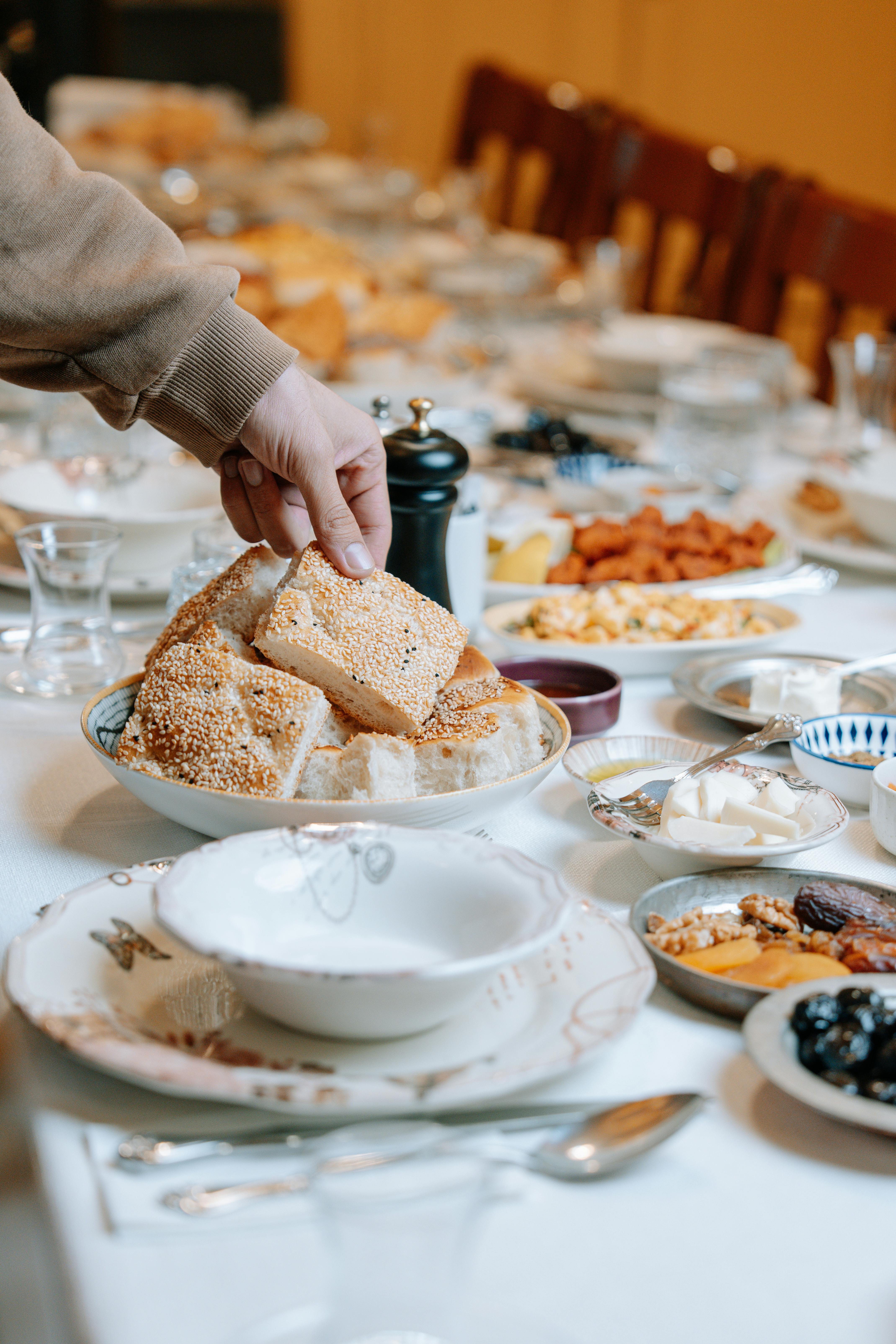 無料 様々な料理が並ぶ豪華な朝食のテーブルで、ゴマパンに手を伸ばす人。 写真素材