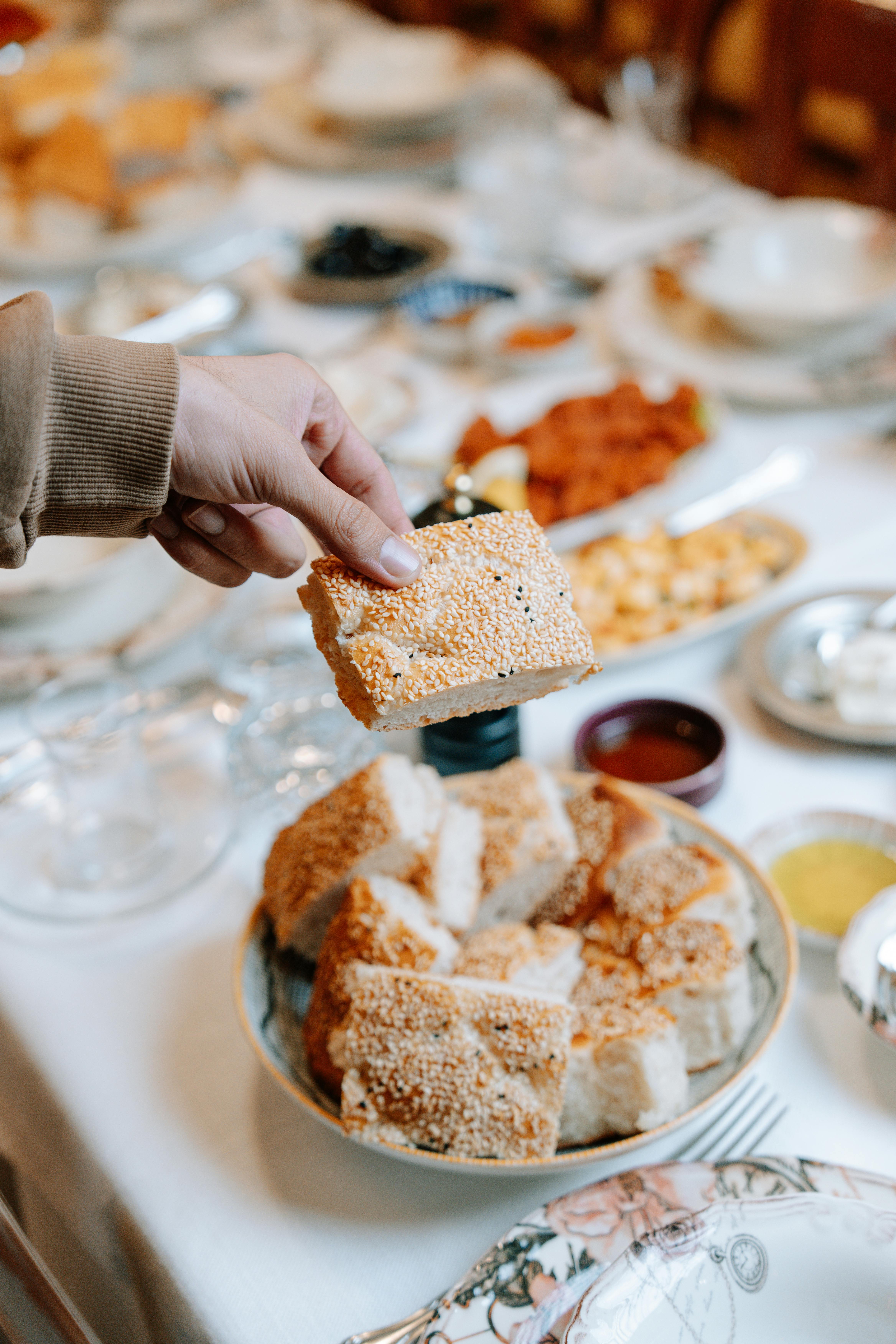 Gratuit Une main tient du pain au sésame au-dessus d'une table dressée avec divers plats, suggérant un repas convivial. Photos