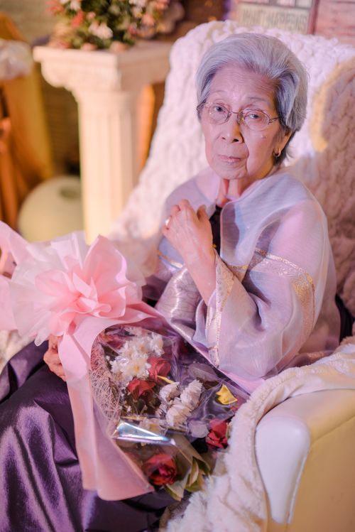Free Graceful senior woman holding a bouquet of flowers, elegantly dressed in a soft setting. Stock Photo