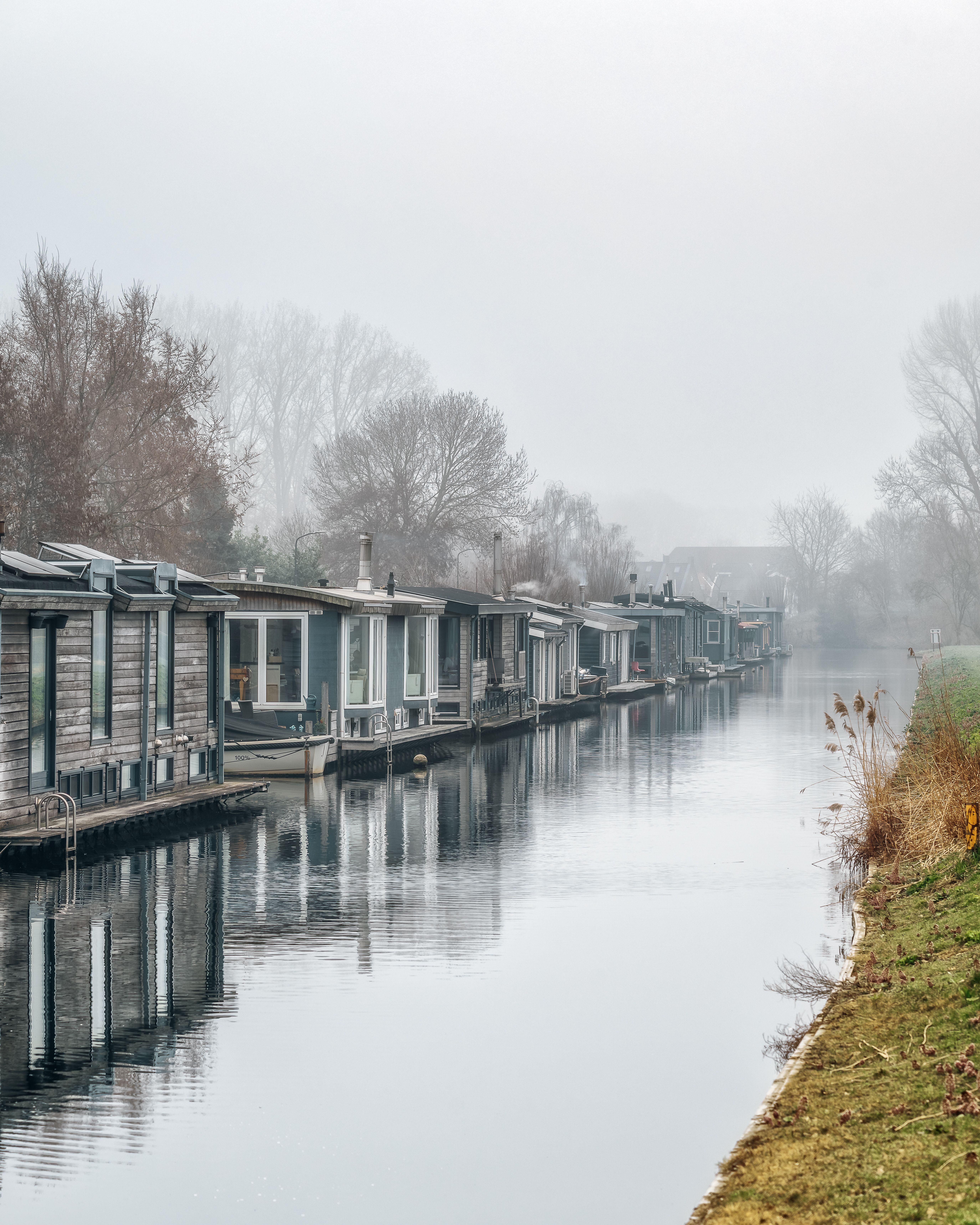 Houseboats lined along a tranquil canal surrounded by misty trees in serene winter ambiance.