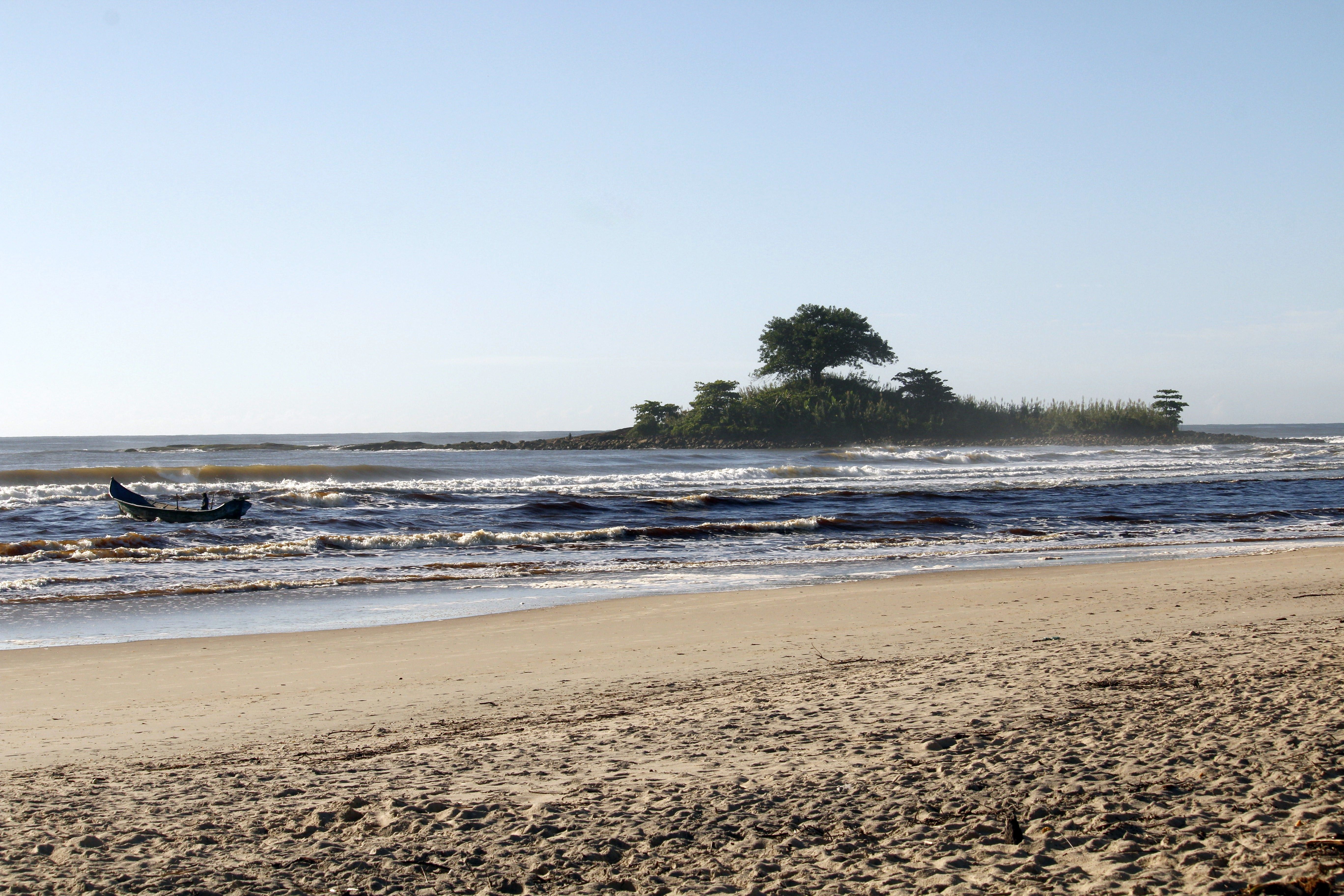 Peaceful beach scene in Guaratuba, Brazil with gentle waves, a boat, and a small island under clear skies.