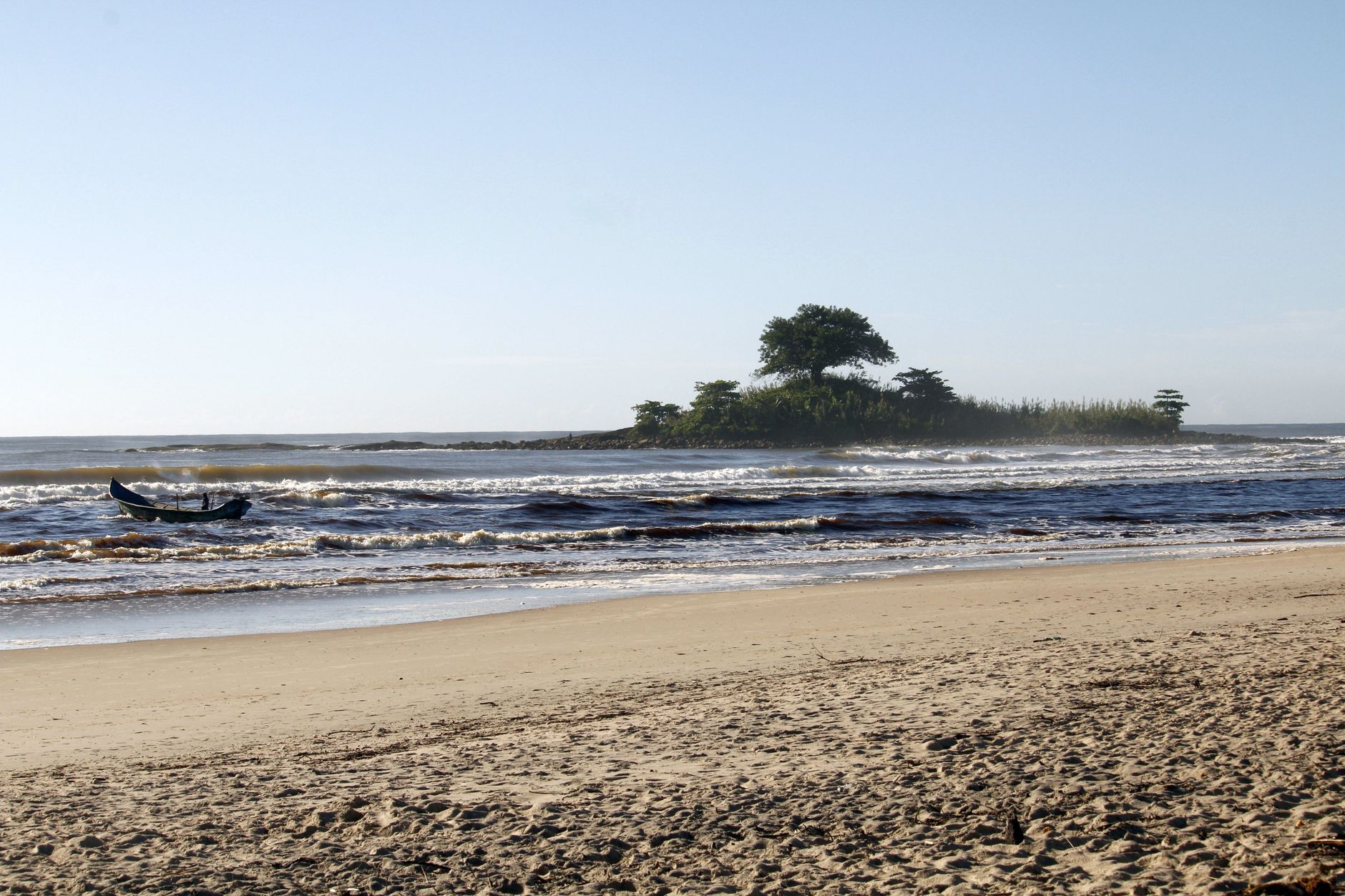 Peaceful beach scene in Guaratuba, Brazil with gentle waves, a boat, and a small island under clear skies.