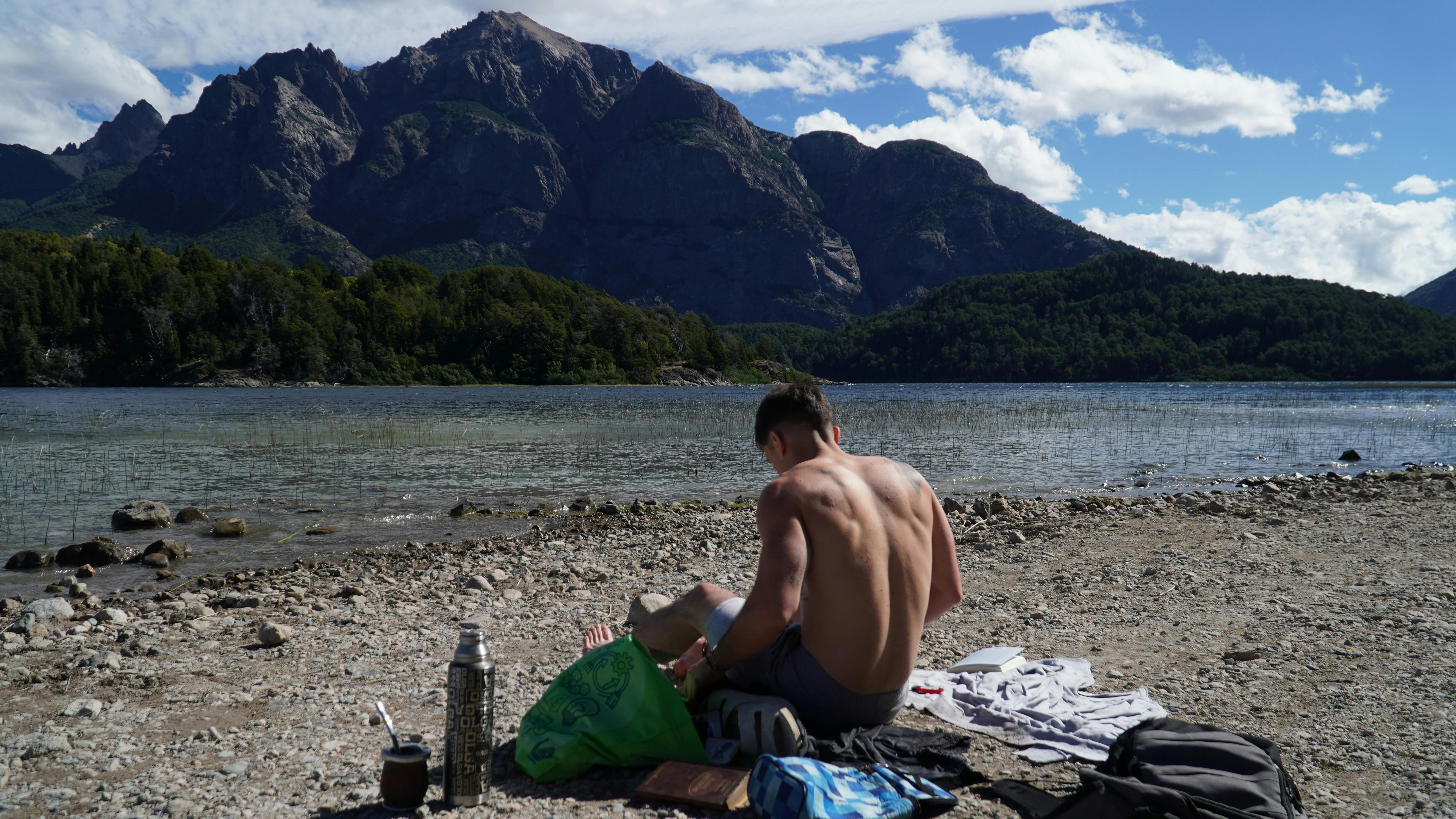 ฟรี ชายคนหนึ่งนั่งอยู่ริมทะเลสาบสีฟ้าใส ล้อมรอบด้วยภูเขาและธรรมชาติอันงดงาม คลังภาพถ่าย