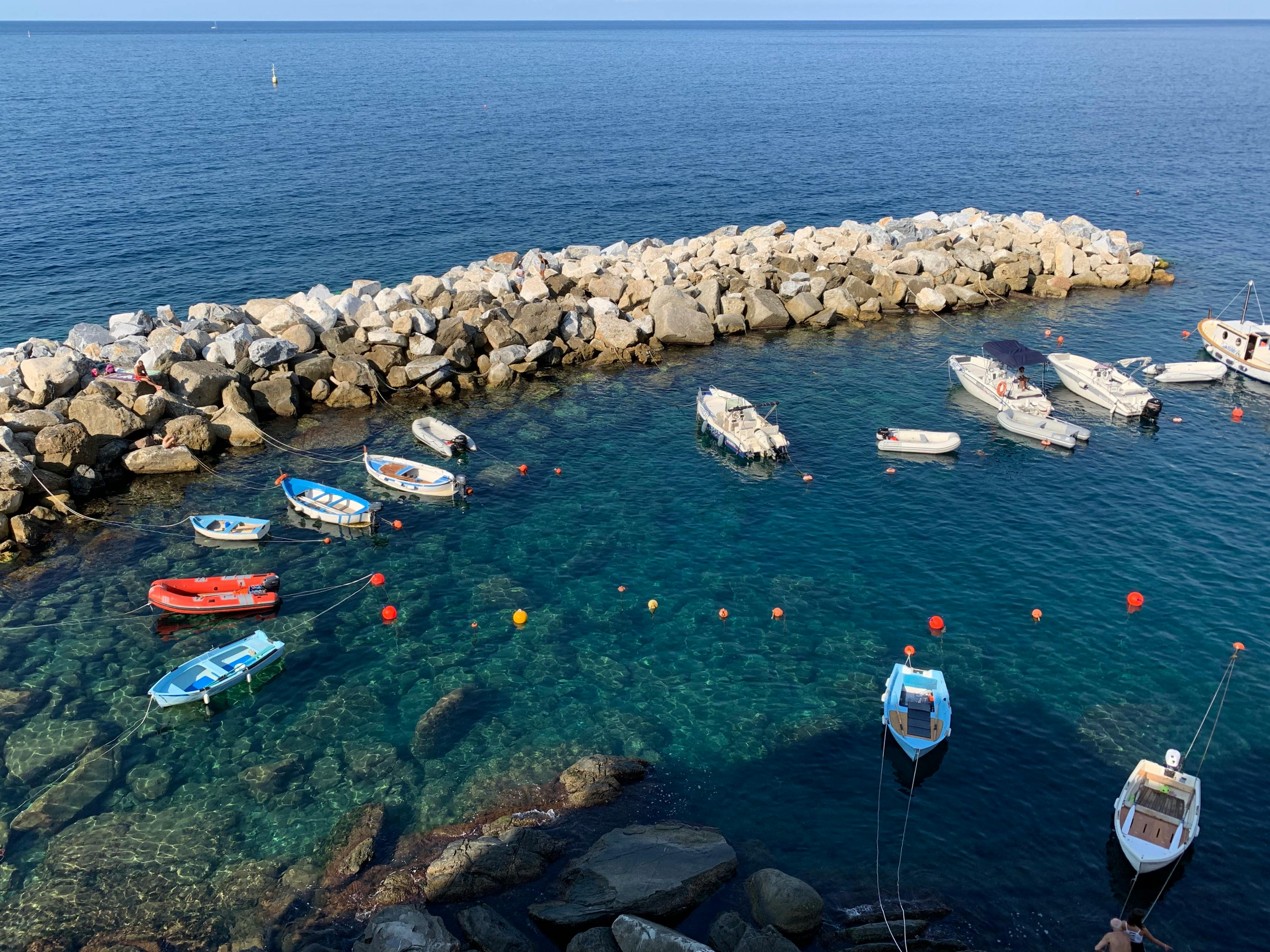 grátis Pequenos barcos coloridos ancorados perto de um quebra-mar rochoso no mar límpido da Ligúria, sob um céu ensolarado. Foto profissional
