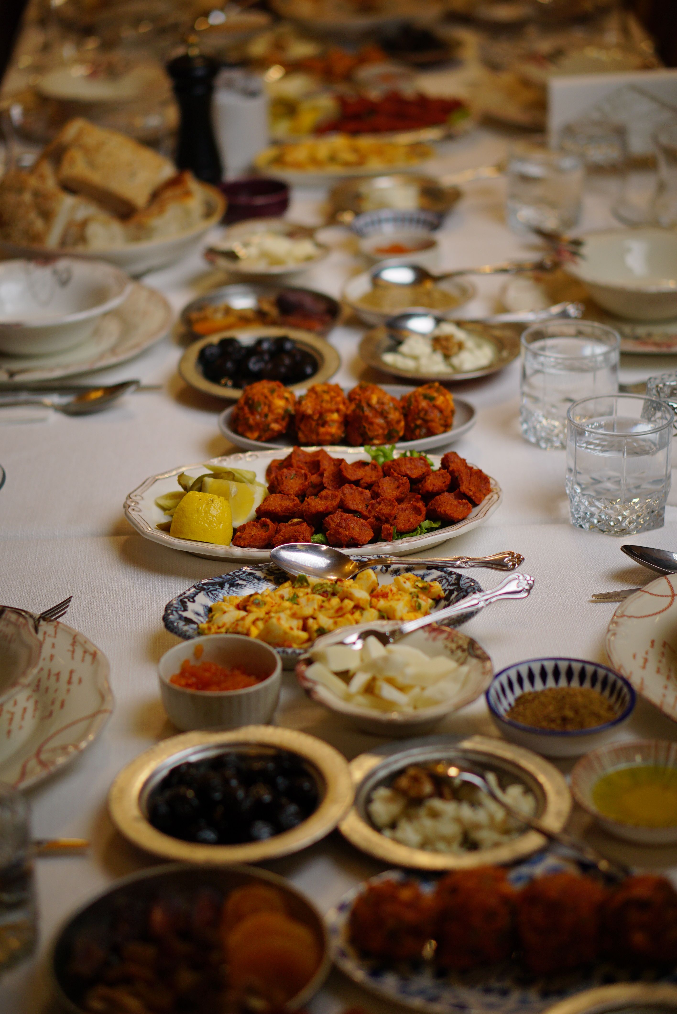 Table dressée à la nappe blanche : plat de boulettes épicées au citron et aux cornichons, beignets de légumes, olives noires, salade d’œufs au paprika et verres à eau taillés