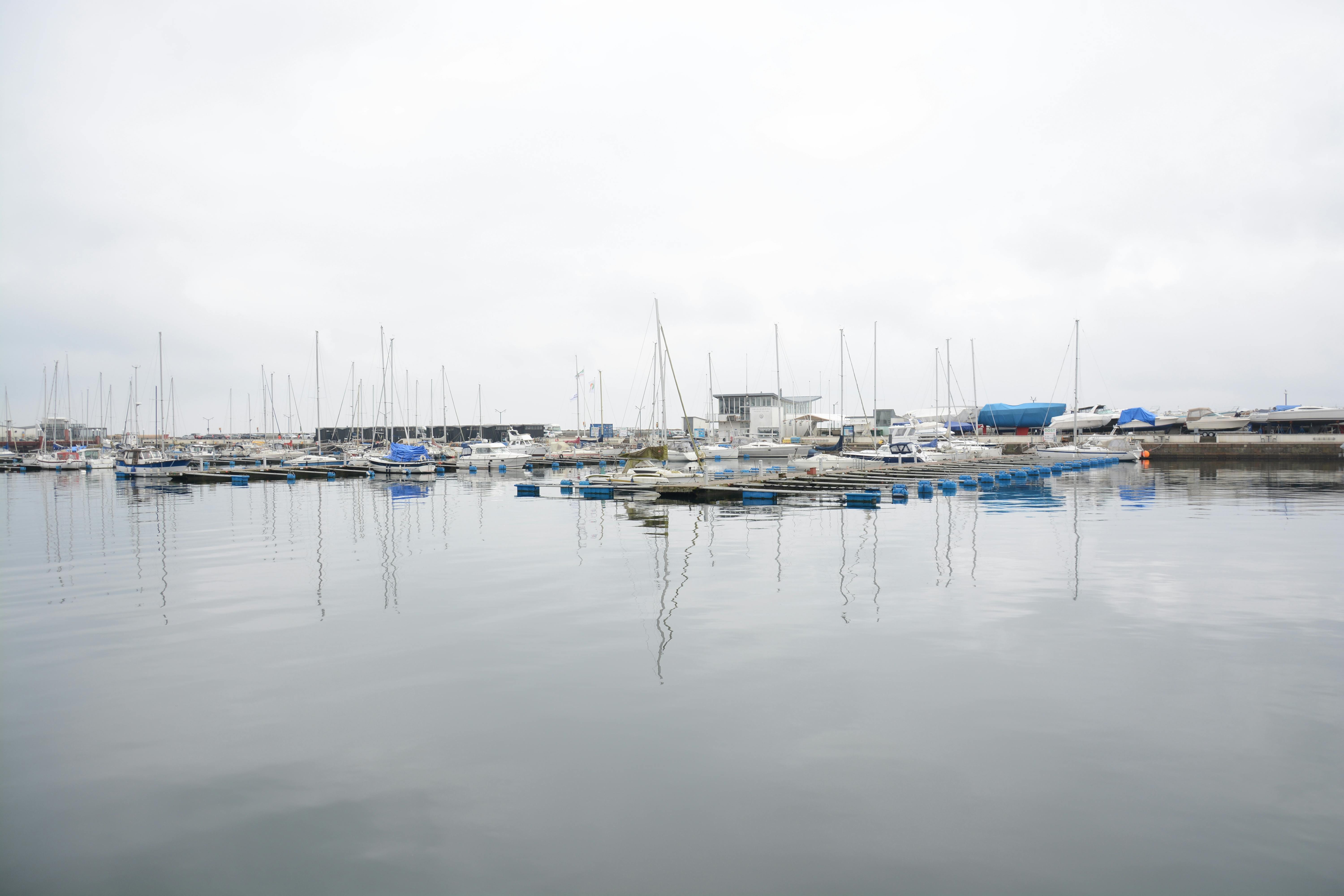 Gratuit Scène paisible de marina avec plusieurs voiliers amarrés sous un ciel couvert se reflétant dans les eaux calmes. Photos