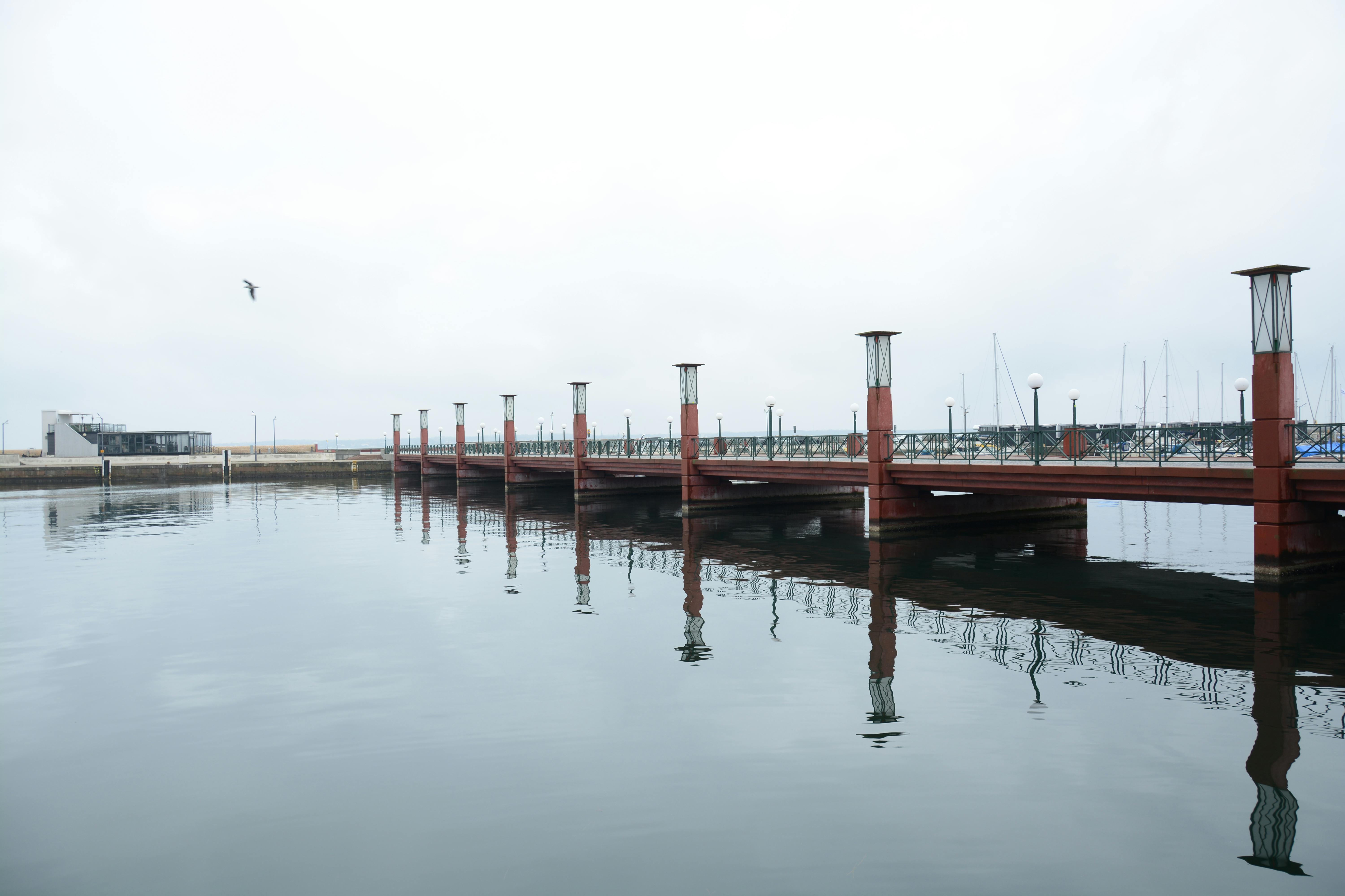 Gratuit Scène portuaire paisible avec un pont rouge se reflétant dans des eaux calmes, sous un ciel couvert. Photos
