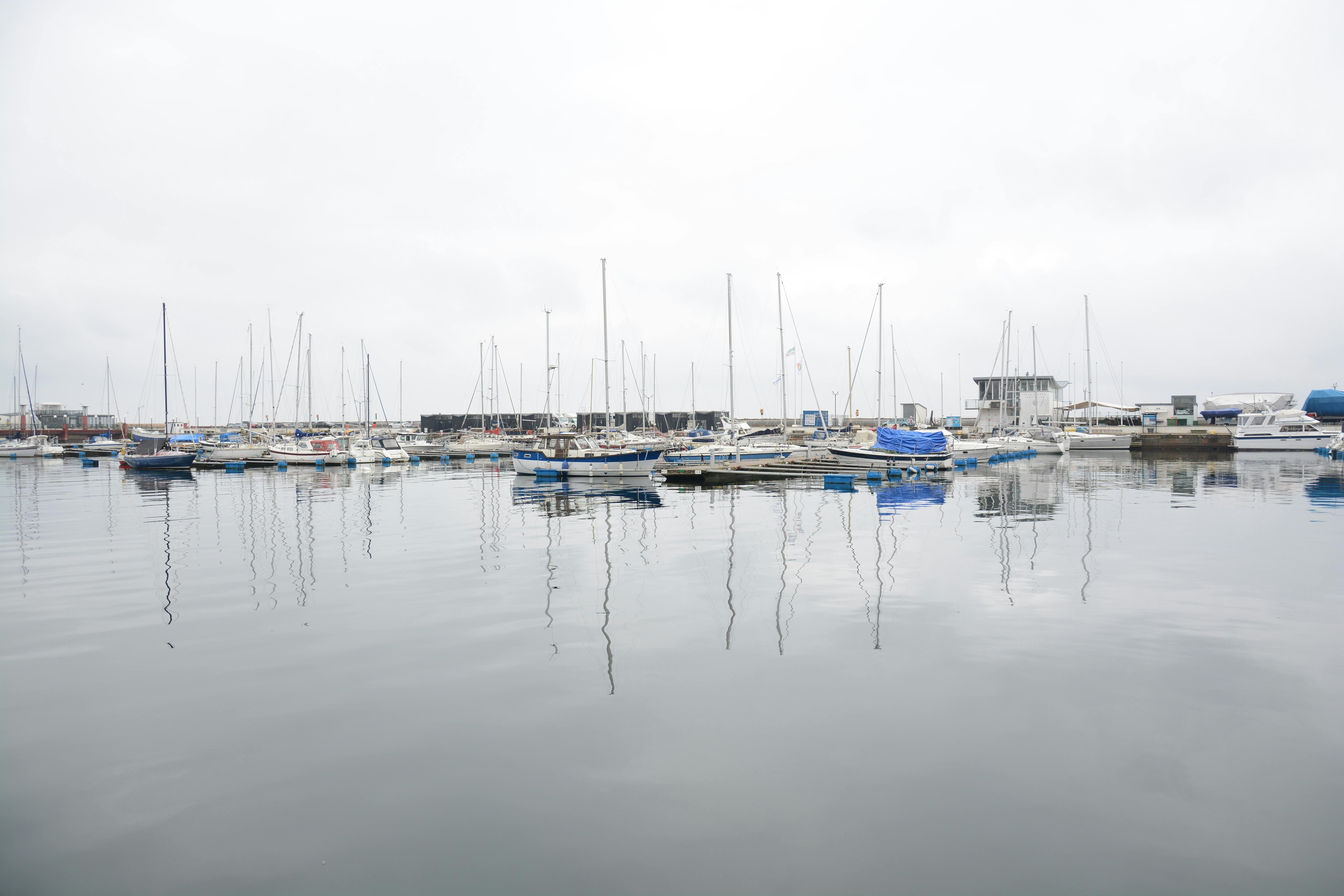 Gratis Rolig marina-scene med fortøyde seilbåter som reflekterer i rolig vann under en overskyet himmel. Arkivbilde