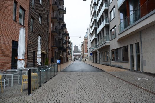 A quiet cobblestone street flanked by modern apartments and outdoor seating in an urban setting.