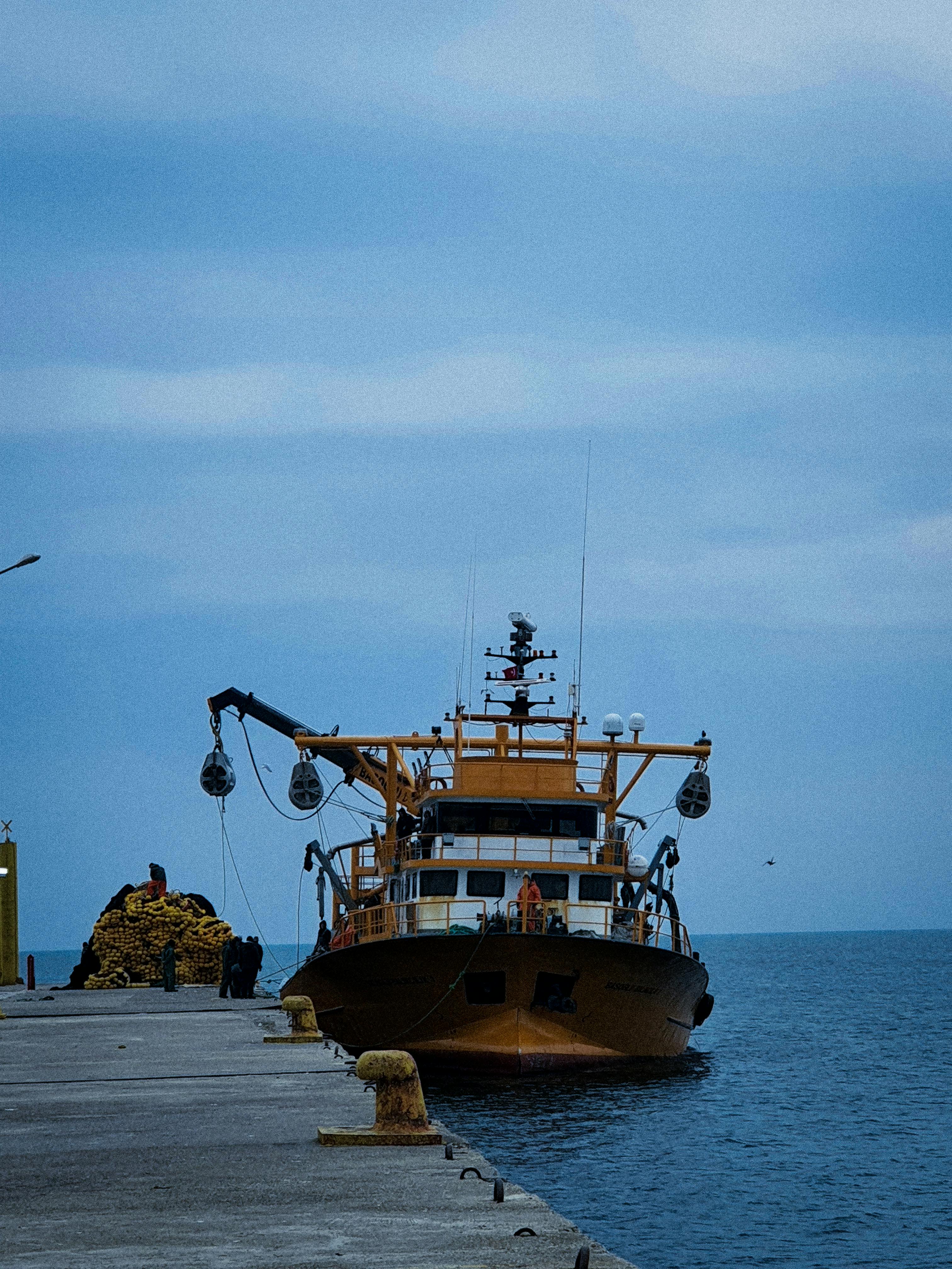 bezplatná Rybářská loď zakotvila u mola pod zataženou modrou oblohou, připravená na dobrodružství na moři. Základová fotografie