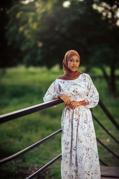 A portrait of a young woman wearing a hijab, standing by a railing outdoors in Nigeria.