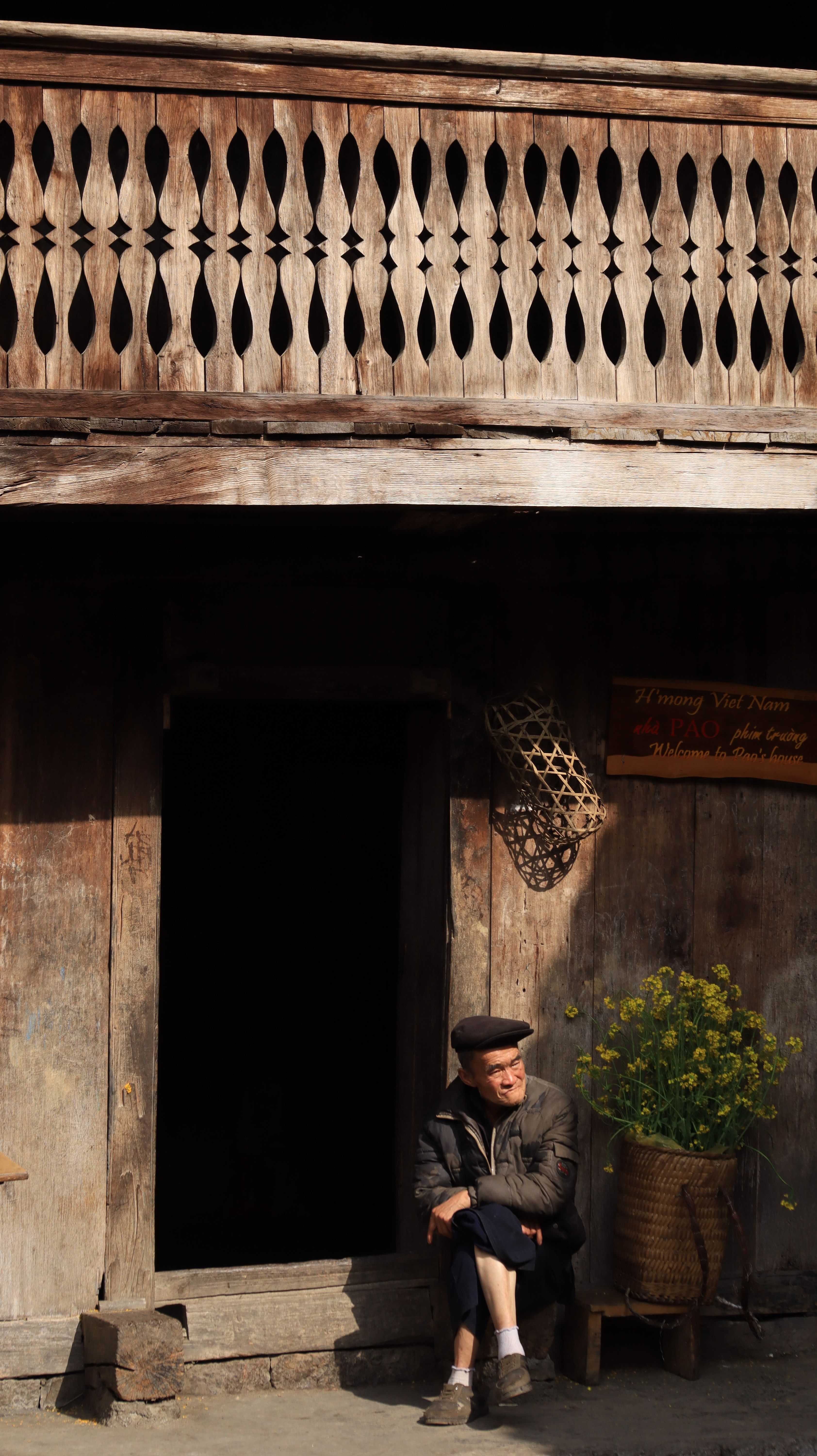 An elderly man sits outside a traditional wooden house in Hà Giang, Vietnam, enjoying the sunlight.