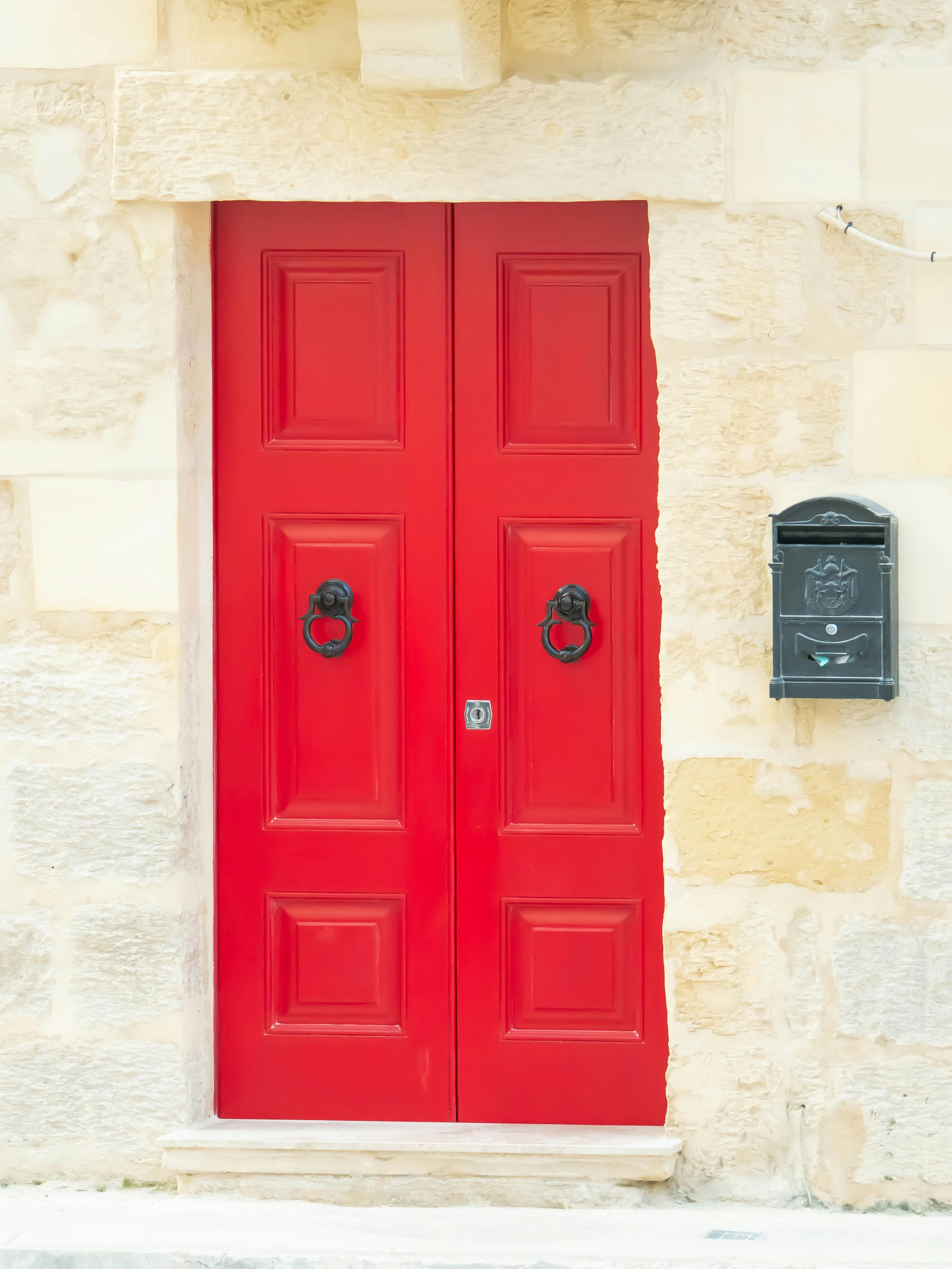Gratis Una llamativa puerta roja con aldabas clásicas, enclavada en una pared de piedra texturizada, junto a un buzón negro. Foto de stock