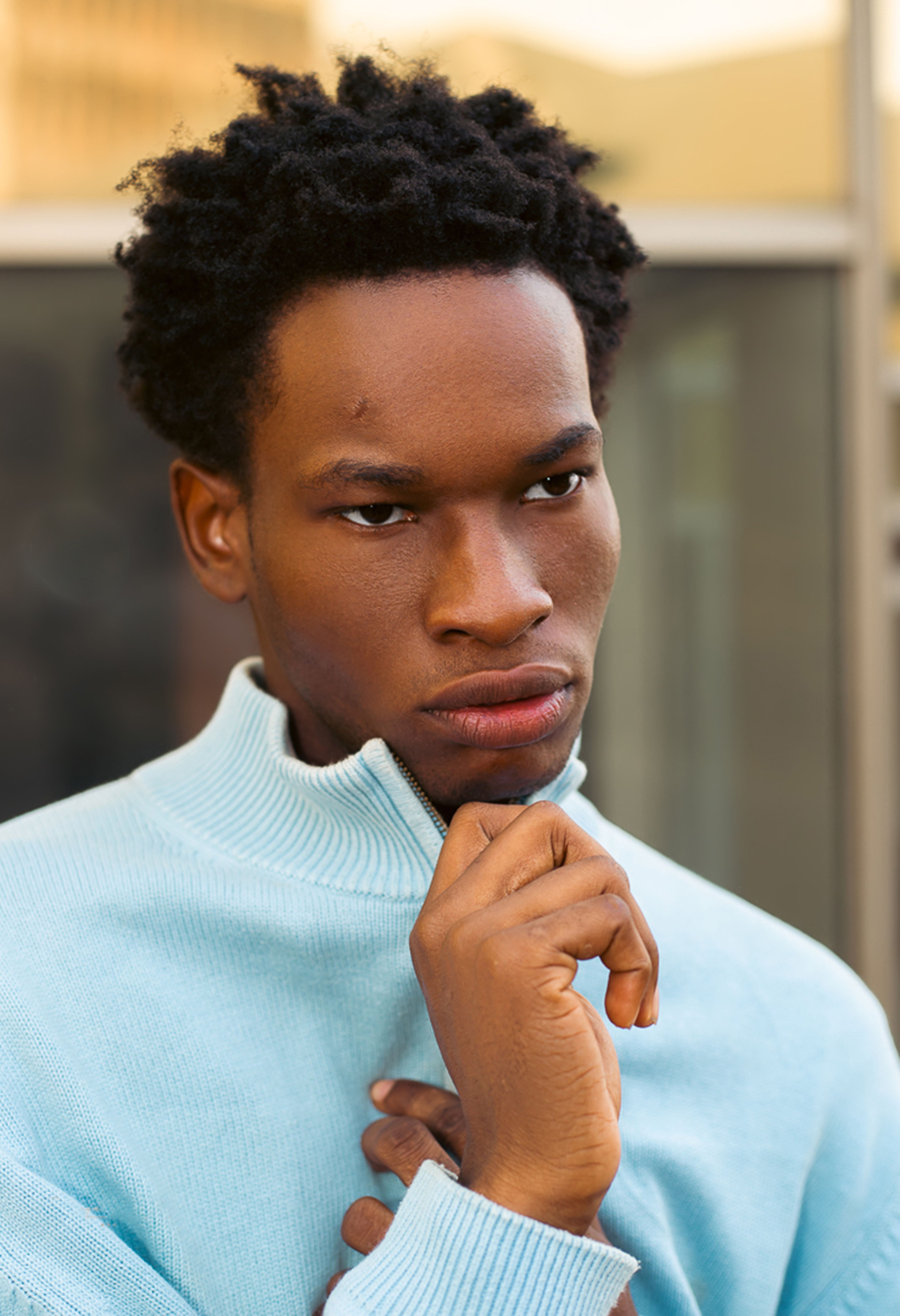 A contemplative person wearing a light blue sweater standing outdoors, in natural light.