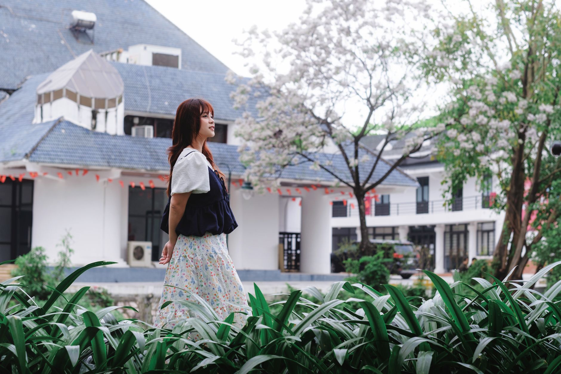A woman strolls peacefully in a lush garden next to a traditional building in Hà Nội, Việt Nam.