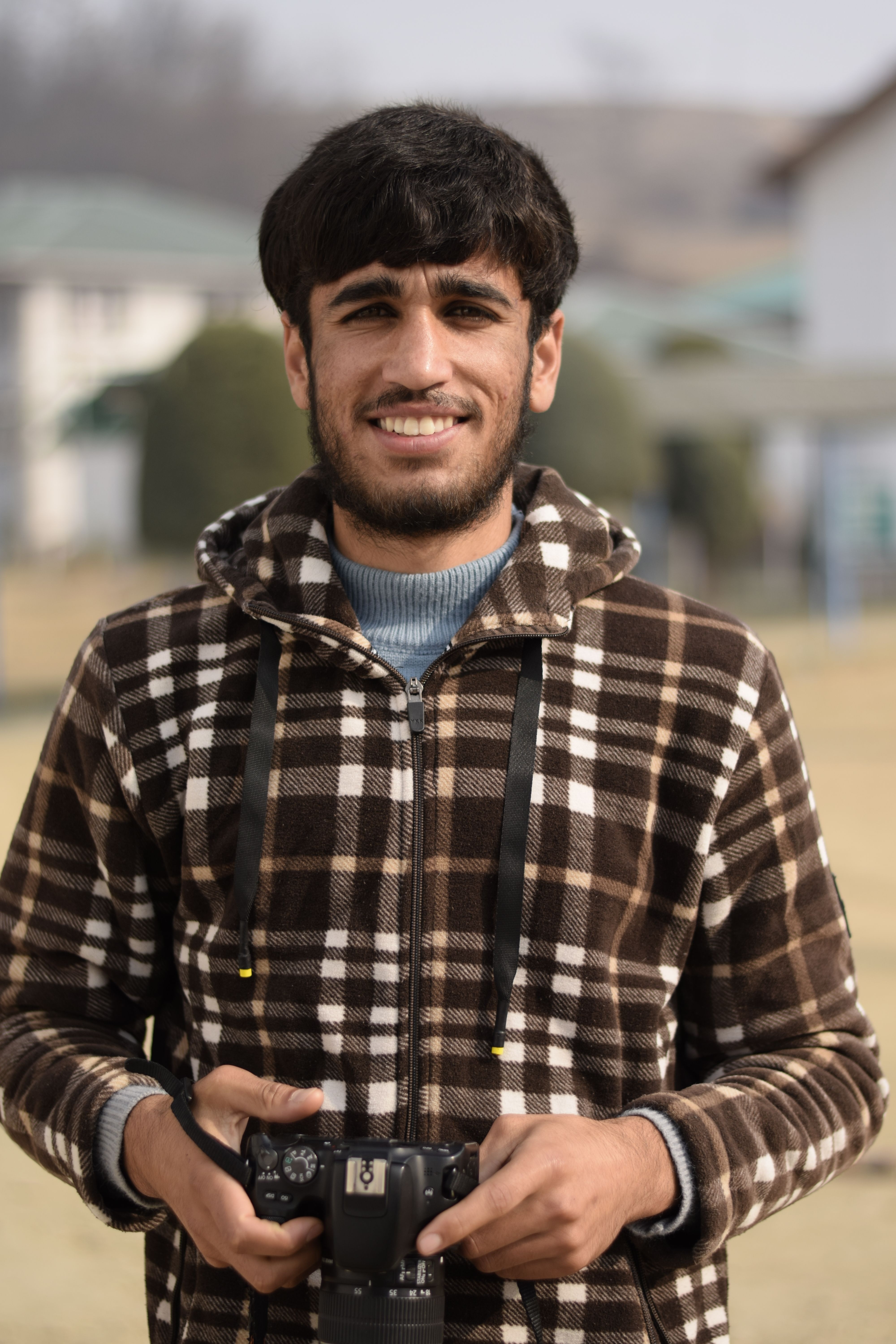 Smiling man in plaid jacket holding camera, standing outdoors during daytime.