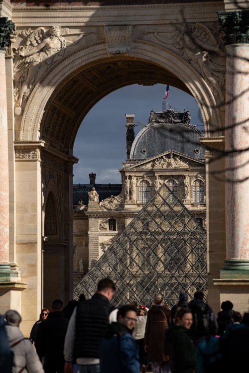 Free Crowds gather at the iconic Louvre Museum entrance, showcasing the pyramid and archway. Stock Photo