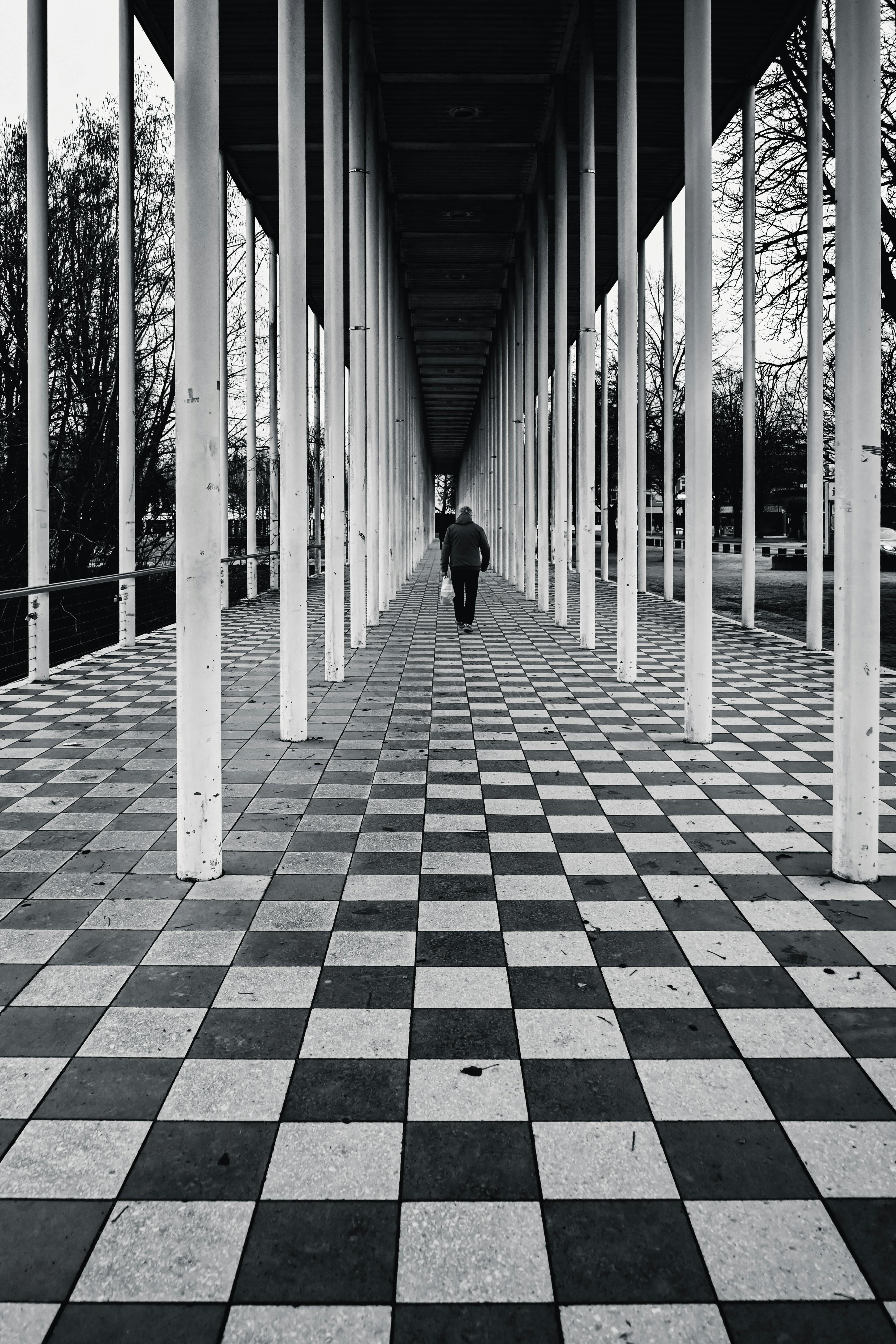 Zadarmo Čiernobiela fotografia osoby kráčajúcej po geometrickom šachovnicovom chodníku so stĺpmi. Fotka z fotobanky