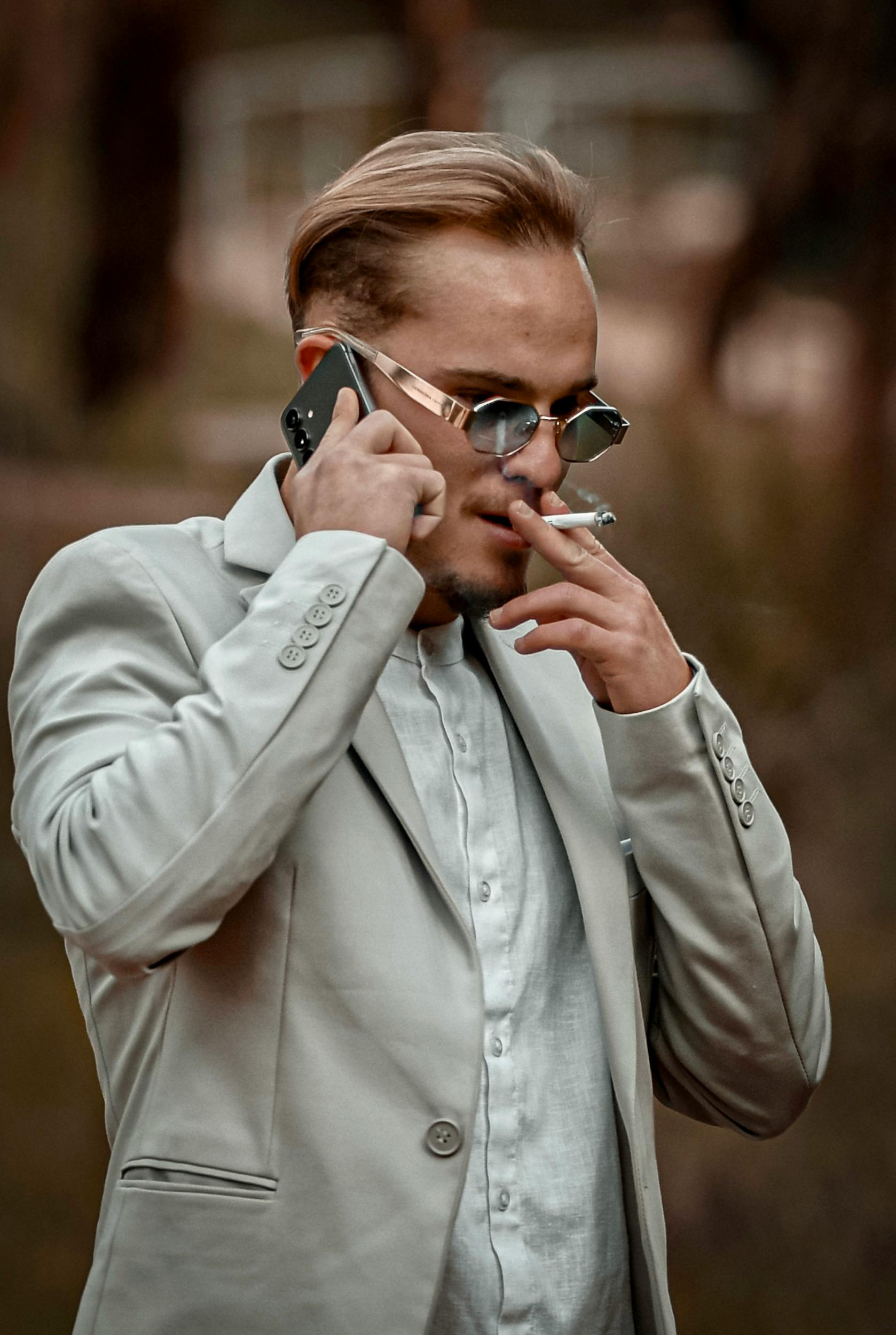 Zadarmo Dobre oblečený muž v slnečných okuliaroch sa rozpráva cez smartfón a fajčí vonku. Fotka z fotobanky