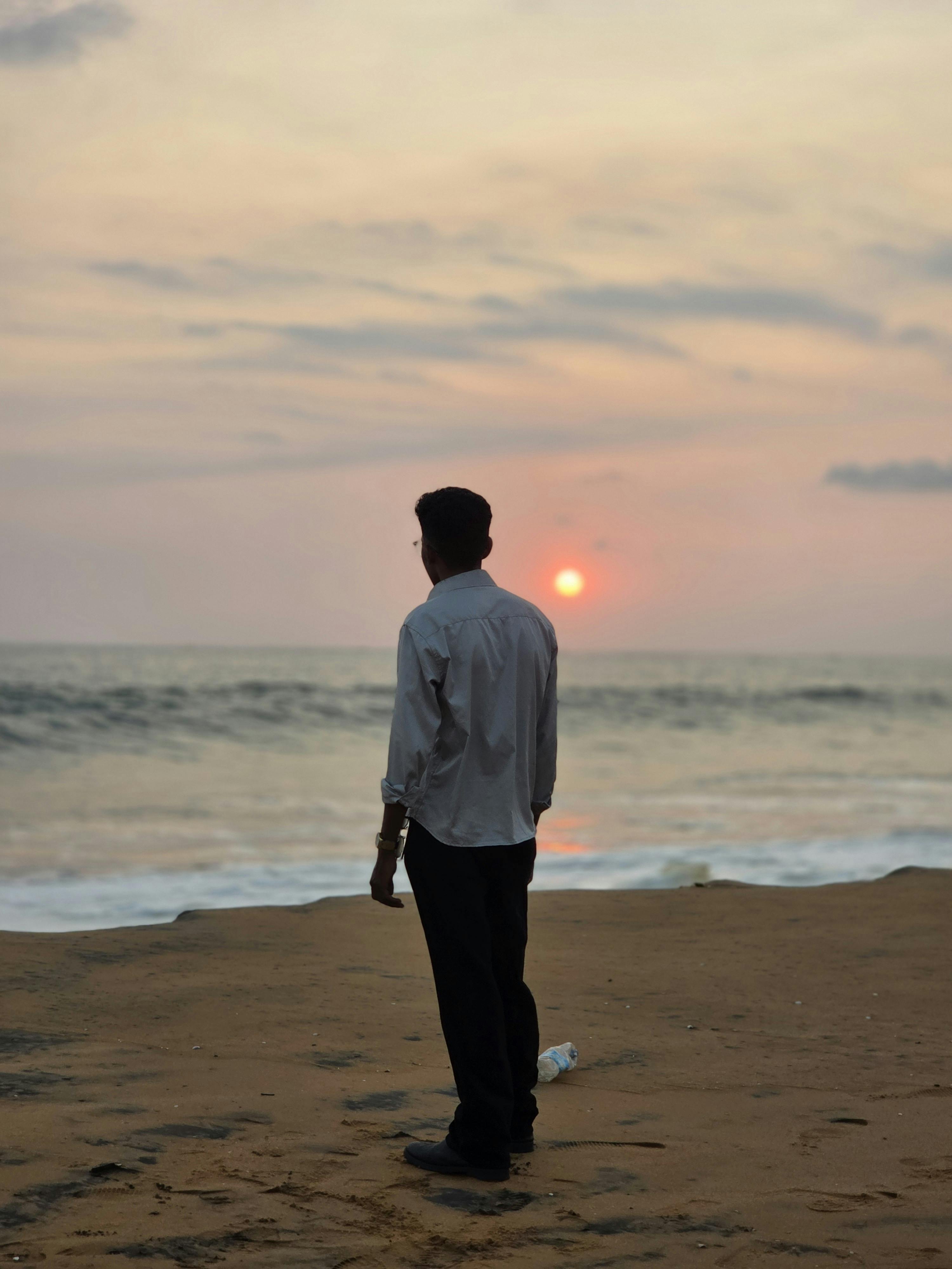 Zadarmo Silueta muža stojaceho na pláži a pozorujúceho pokojný západ slnka nad oceánom. Fotka z fotobanky