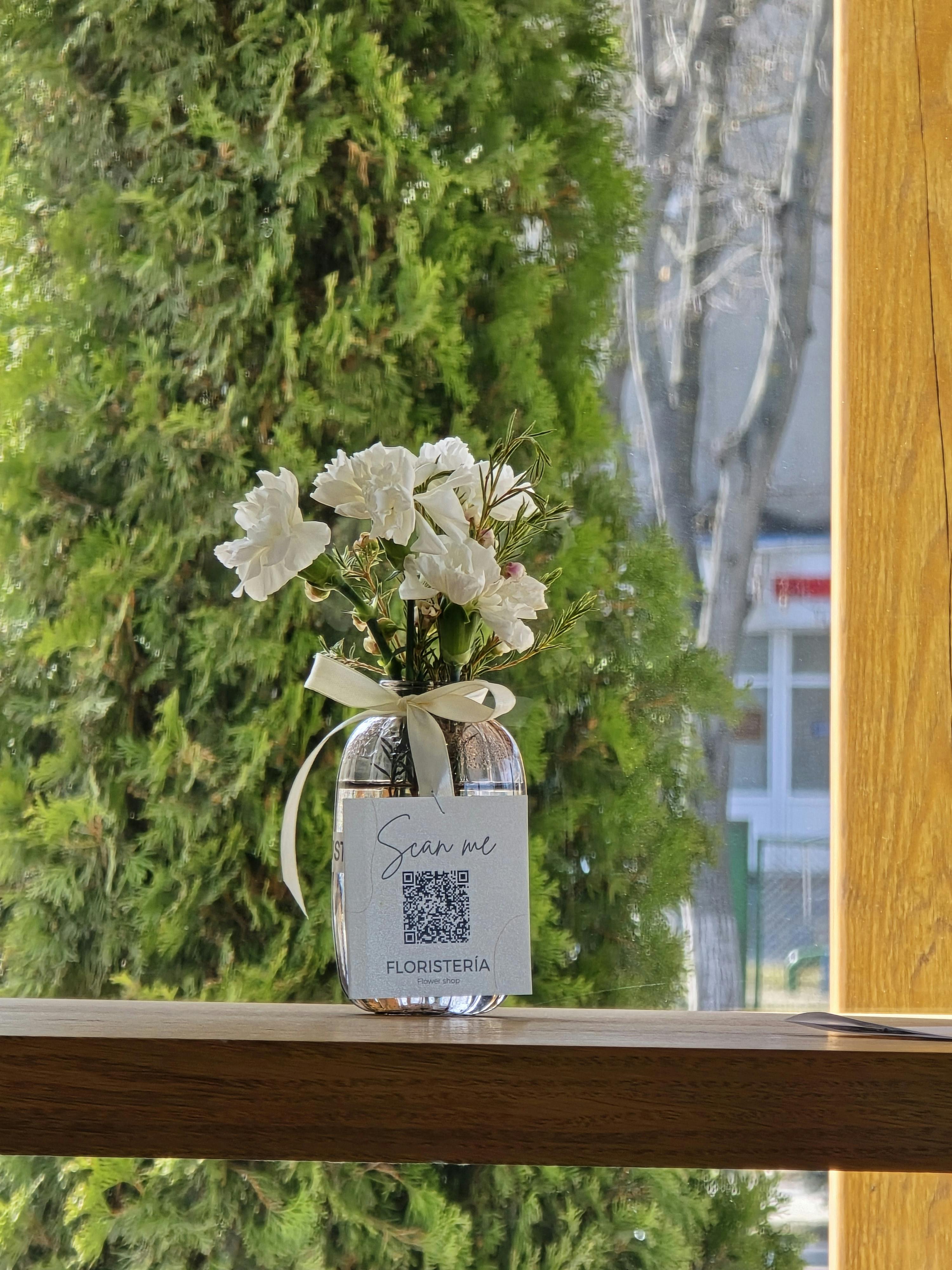 Zadarmo Štýlová sklenená váza s bielymi kvetmi a QR kódom, umiestnená na parapete s výhľadom von. Fotka z fotobanky