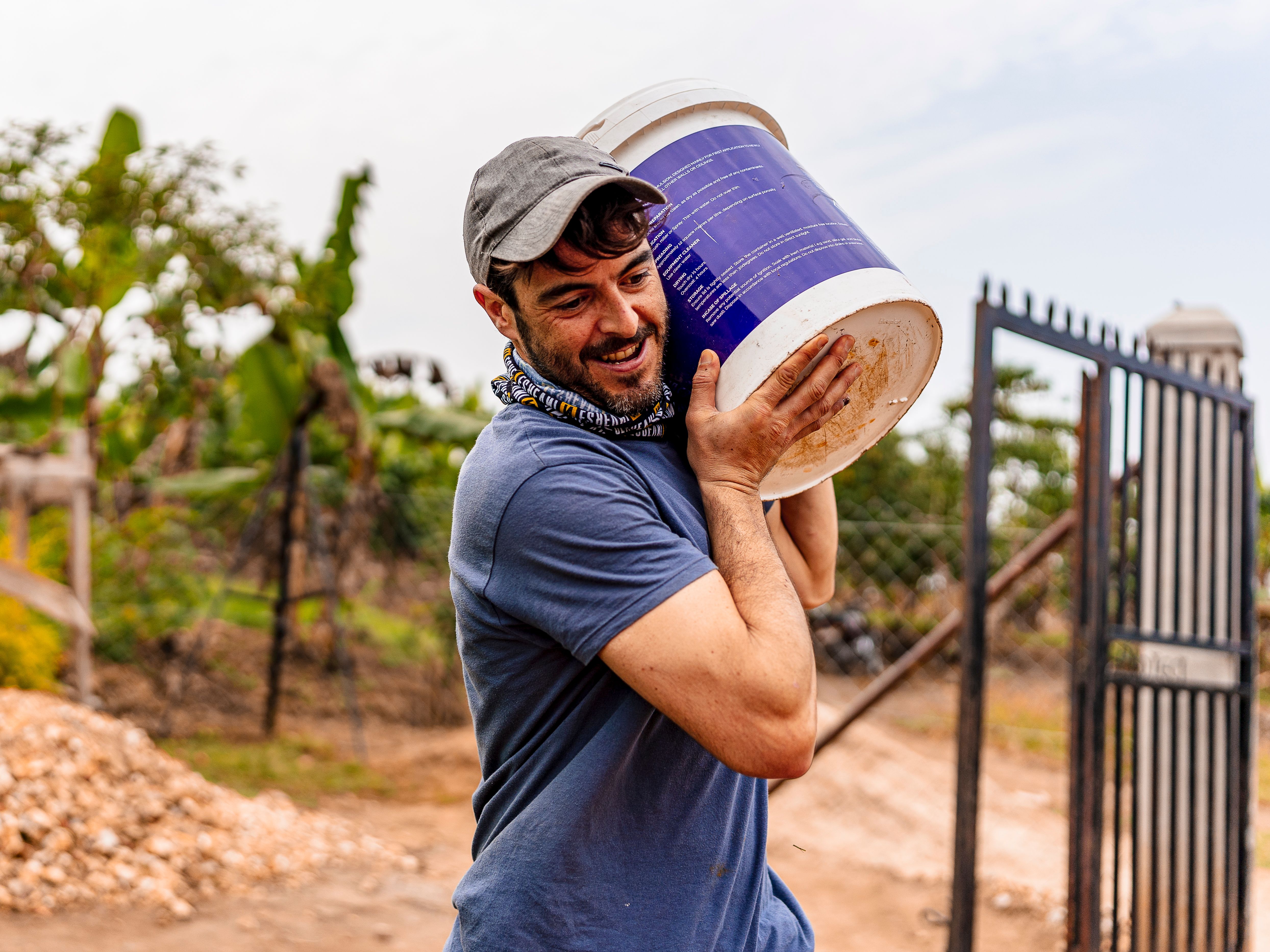 A smiling man in casual clothing carries a large blue bucket outdoors on a sunny day.